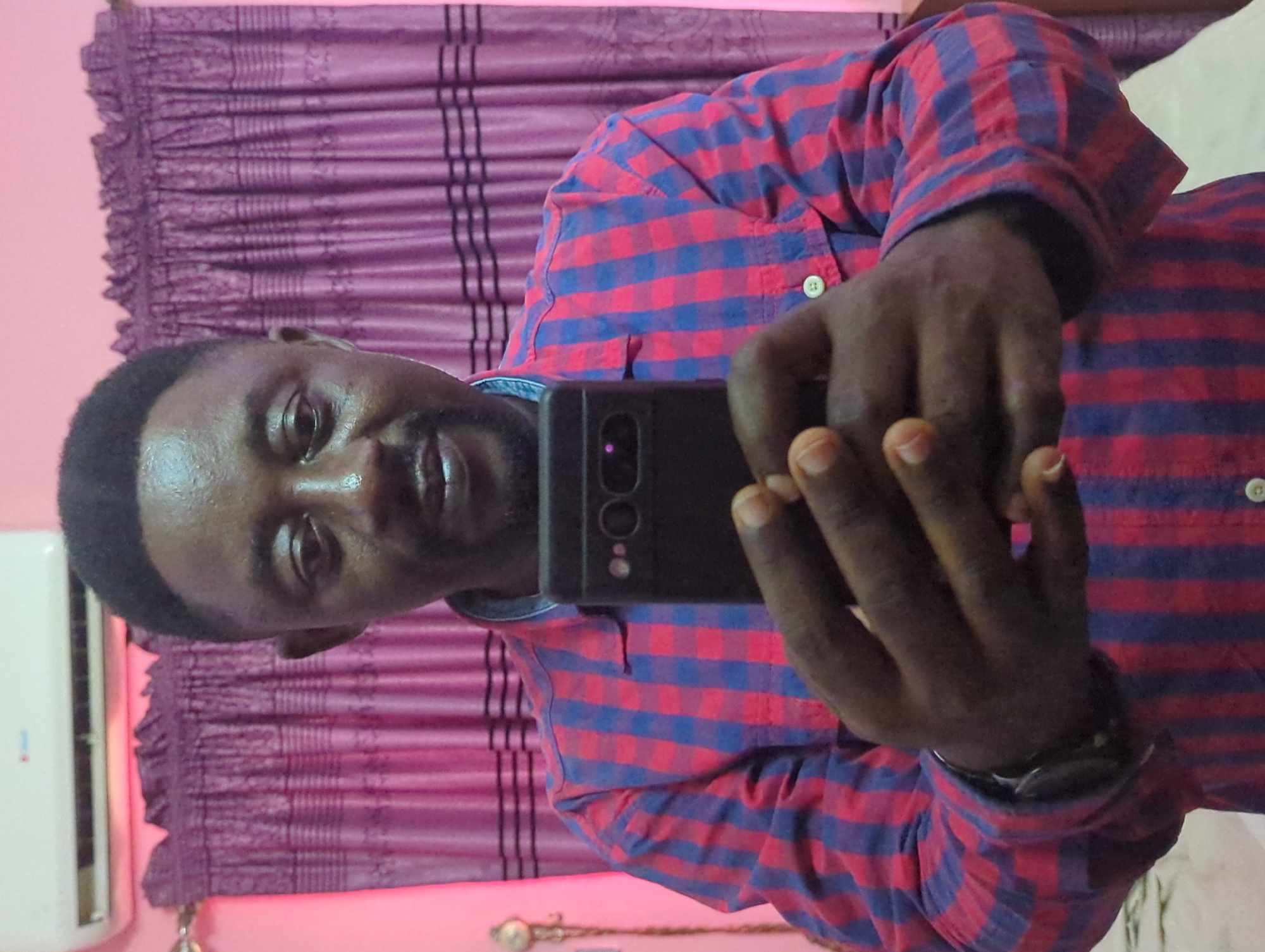 A man is taking a selfie in an indoor setting. He is wearing a red and blue checkered shirt. He holds a dark-colored smartphone in his hands, positioning it to capture his image. The background consists of a pink wall and a maroon curtain with decorative elements. An air conditioning unit is mounted on the wall to the left. The location is in the city of Unknown, Unknown.