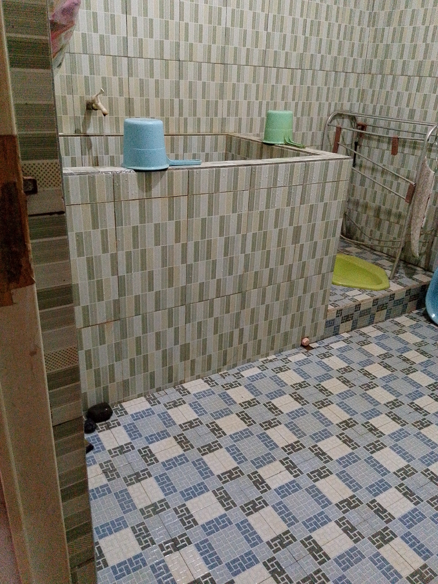 This is an indoor image of a small bathroom in Babadan, Indonesia. The bathroom features light green and white tiled walls and a patterned tile floor in shades of blue, white, and gray. A raised tiled structure, likely a built-in bathtub or shower base, is present, with a light blue plastic bucket with a handle on top. A green plastic bucket is also visible on the same structure. A metal drying rack with several items hanging on it is positioned to the right. A light green squat toilet or bidet is on a tiled platform to the right of the main tiled structure. A water faucet is visible on the tiled wall above the blue bucket. Two dark objects, possibly slippers or mats, are on the floor near the left edge of the image. A small, reddish-brown spherical object is on the floor near the base of the tiled structure. The overall condition of the bathroom appears used but functional.