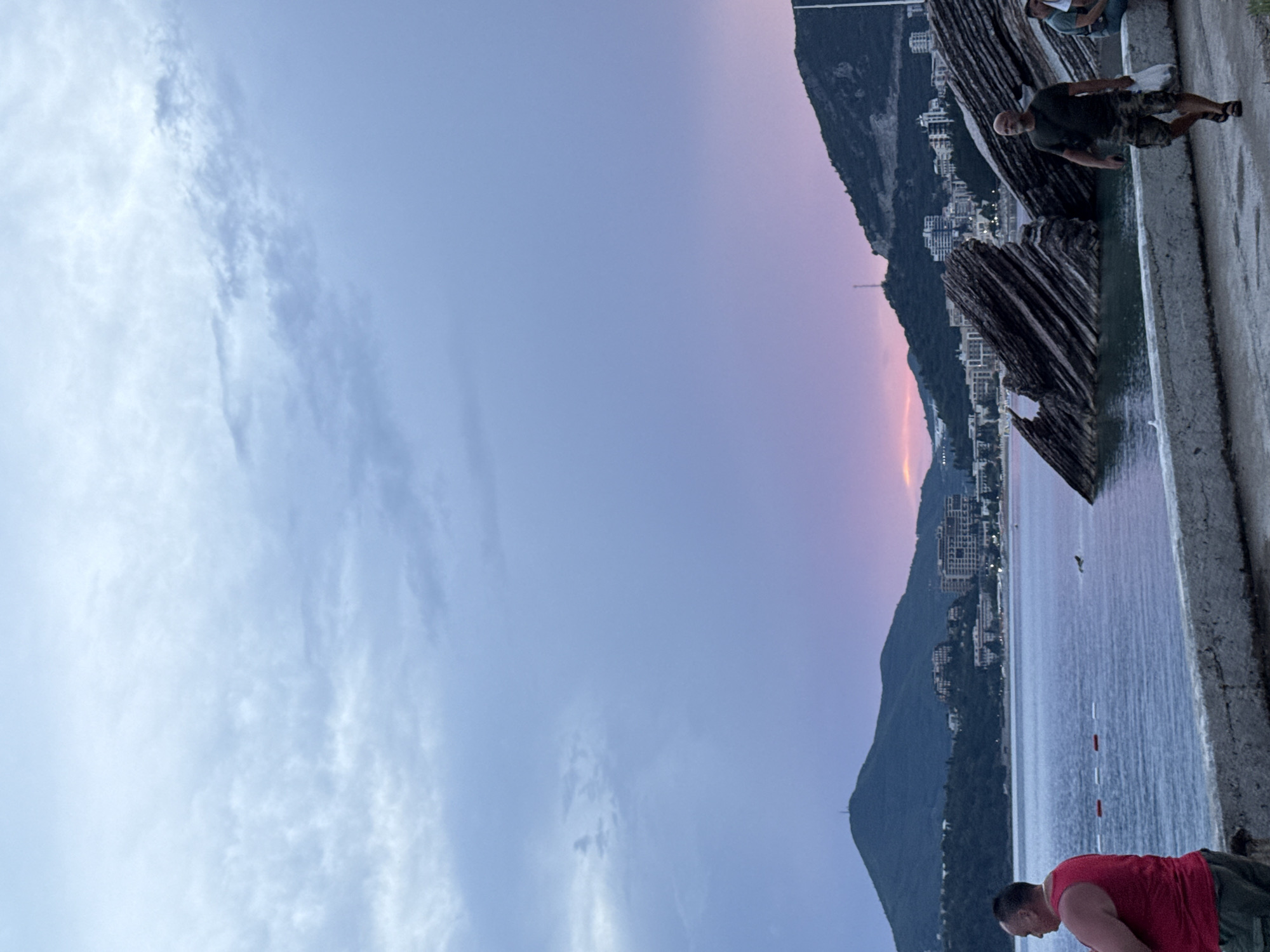 The image captures a serene evening scene in Rafailovići, Montenegro. The sky is a canvas of soft hues, transitioning from a pale blue on the left to a delicate blend of pink and purple on the right, indicating sunset or twilight. Wispy clouds drift across the sky, adding texture to the twilight atmosphere.
The foreground features a concrete walkway or embankment, with a rough texture and a slight sheen of water. To the right of the walkway, the calm sea reflects the colors of the sky. In the water, a few red and white buoys are visible, marking a swimming area.
On the left side of the image, a lush green mountain slopes down towards the sea. Nestled on the mountainside is a cluster of buildings, appearing to be hotels or apartments, their lights beginning to twinkle as the day fades. The architecture suggests a coastal resort town.
Several people are present. In the lower right, a man in a red tank top and dark shorts is captured in a bent-over posture, possibly resting or engaged in an activity near the water's edge. Further up the walkway, another man in a dark t-shirt and camouflage shorts stands, looking towards the sea and the distant landscape. A partial view of a third person can be seen in the upper right, near the edge of the frame.
Dominating the mid-ground are several large, abstract, wooden structures, with a rough, layered texture, resembling driftwood or natural formations. These structures add a unique artistic or natural element to the scene.
The overall mood is peaceful and contemplative, with the natural beauty of the landscape enhanced by the soft evening light and the presence of human activity. No visible text is present in the image.