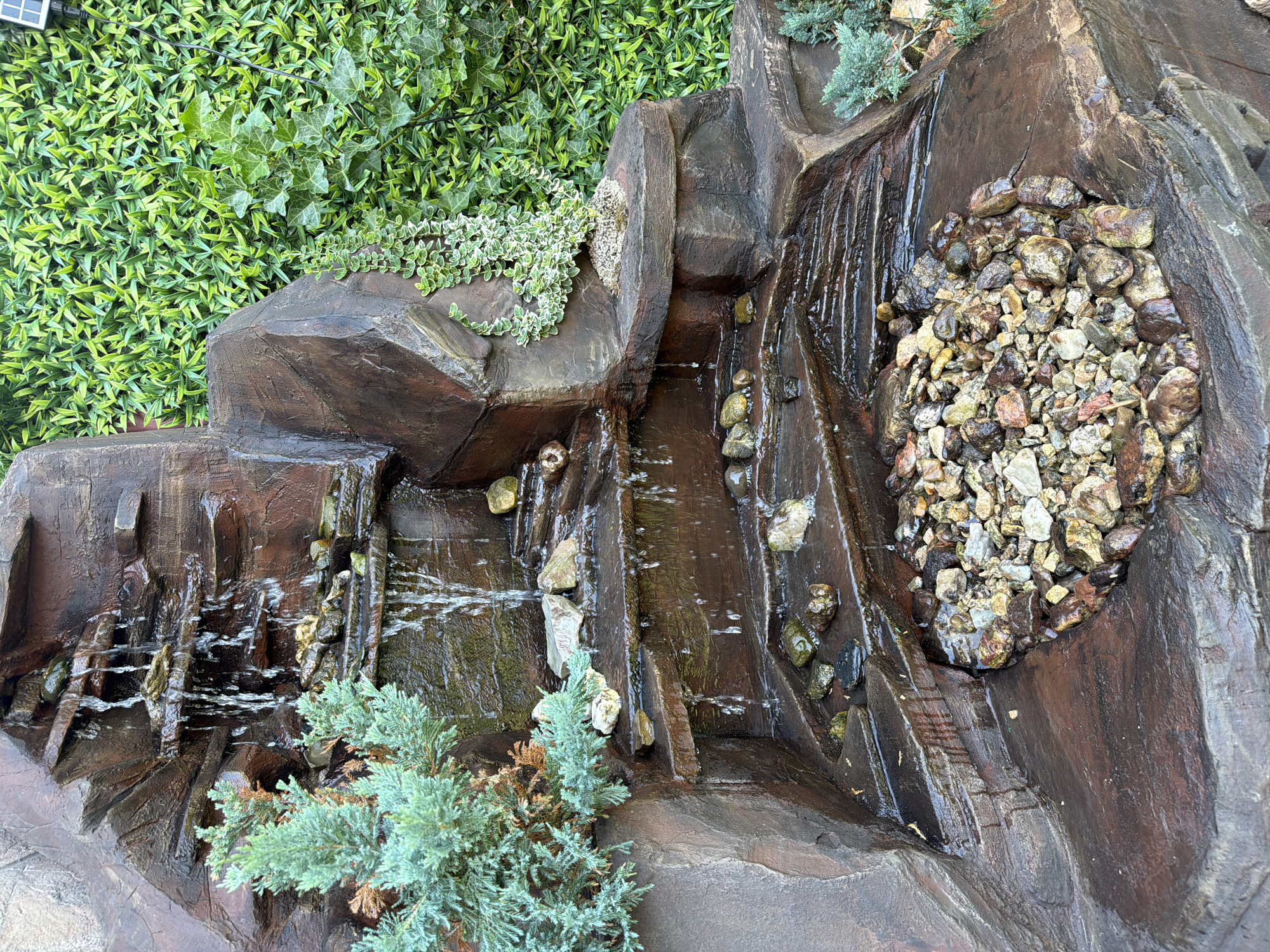 An artificial rock waterfall is shown with water flowing down multiple tiers. The rocks are reddish-brown and textured, with some appearing darker and wet. Small green bushes are planted on some of the rock ledges, and a cluster of small stones is at the base of the waterfall. Behind the waterfall, a dense green wall of artificial foliage is visible. The location is described as Unknown, Unknown.