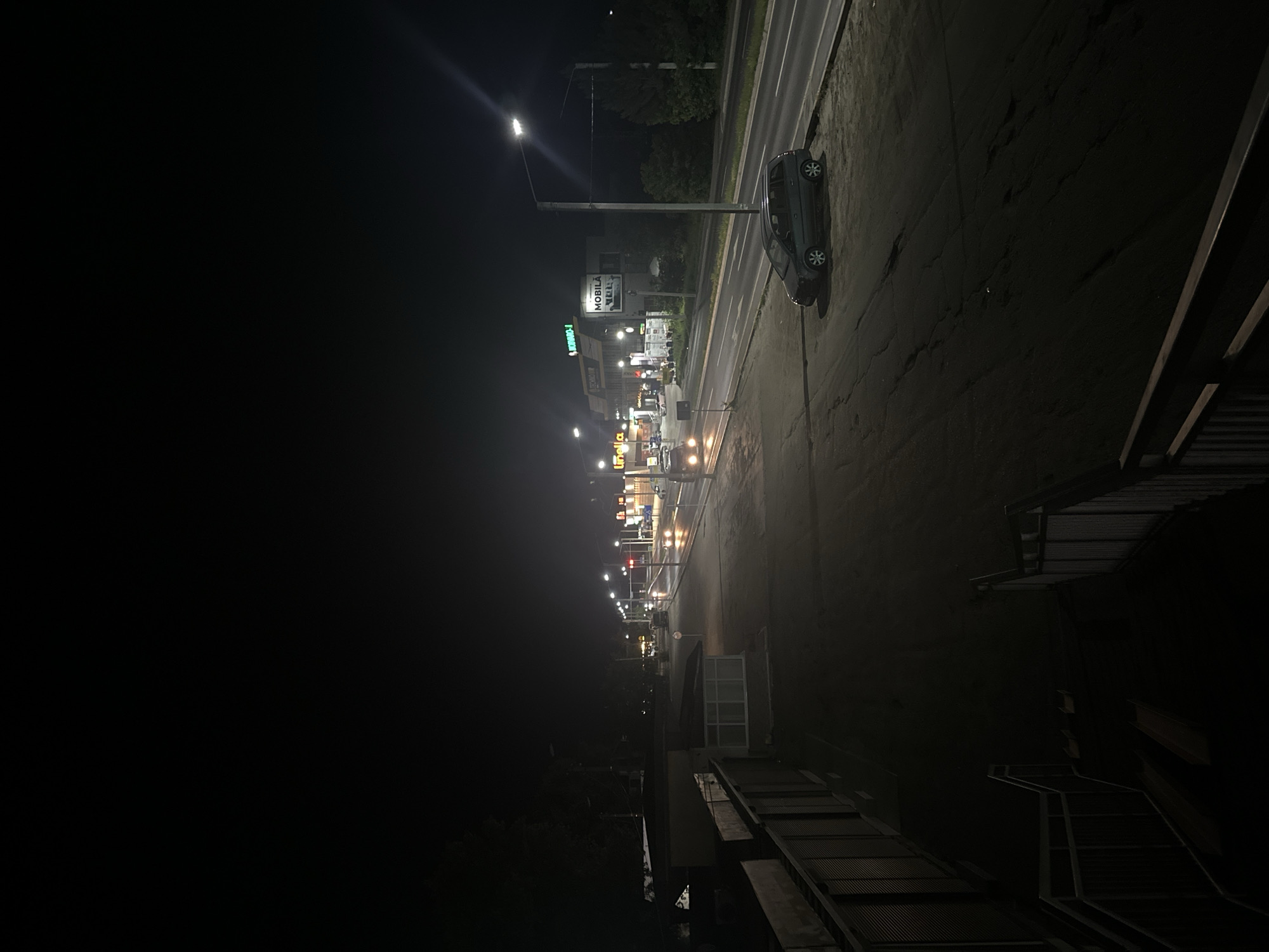 The image depicts a nighttime street scene in Strășeni, Moldova. The foreground shows a dark, paved area with a grey, four-door hatchback car parked on the right side. A metal fence and stairs are visible on the left. The midground features a road with visible lane markings, illuminated by streetlights and the lights from businesses on the right. Several cars are in motion on the road, their headlights and taillights creating streaks of light. A few buildings with illuminated signage are present along the roadside, including one that appears to be a technology store. The background consists of dark trees and foliage, suggesting a less developed area beyond the immediate urban environment. The overall atmosphere is one of a quiet, somewhat desolate, urban fringe at night.