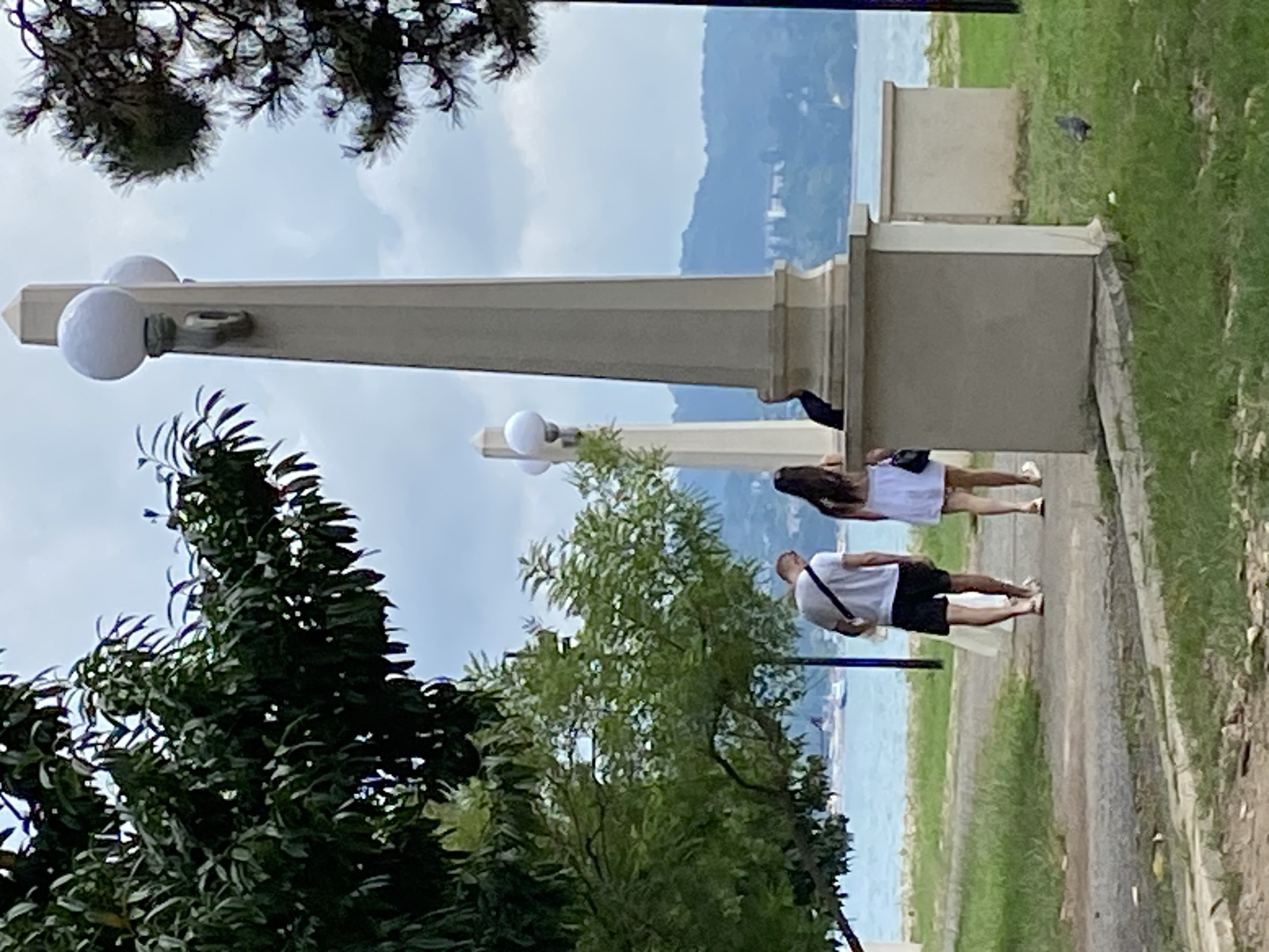 A couple walks away from the camera on a paved path in a park setting. The man on the left wears a white t-shirt and black shorts, carrying a white bag over his left shoulder. The woman on the right wears a white dress and carries a black purse on her right shoulder. They are walking towards a body of water, likely the Black Sea, which is visible in the background. Beyond the water, a hazy coastline with buildings and hills can be seen. A tall obelisk-shaped lamppost with two spherical lights is prominent in the foreground to the right of the couple. Lush green trees with dark foliage flank the path and the lamppost. The ground on either side of the path is covered in grass, with some scattered debris. The sky is overcast. This scene is located in Sokhumi, Abkhazia, likely in a park along the coastline.
