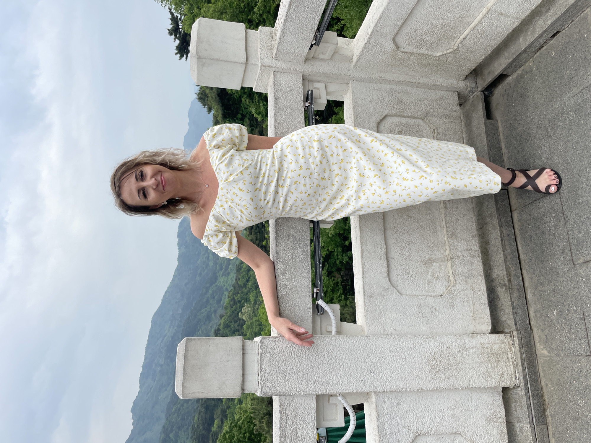 A woman is leaning against a white stone railing, wearing a white dress with a small yellow floral pattern and dark brown sandals. She has blonde hair. Behind her is a forested mountain range under an overcast sky. The location is Huairou District, China.