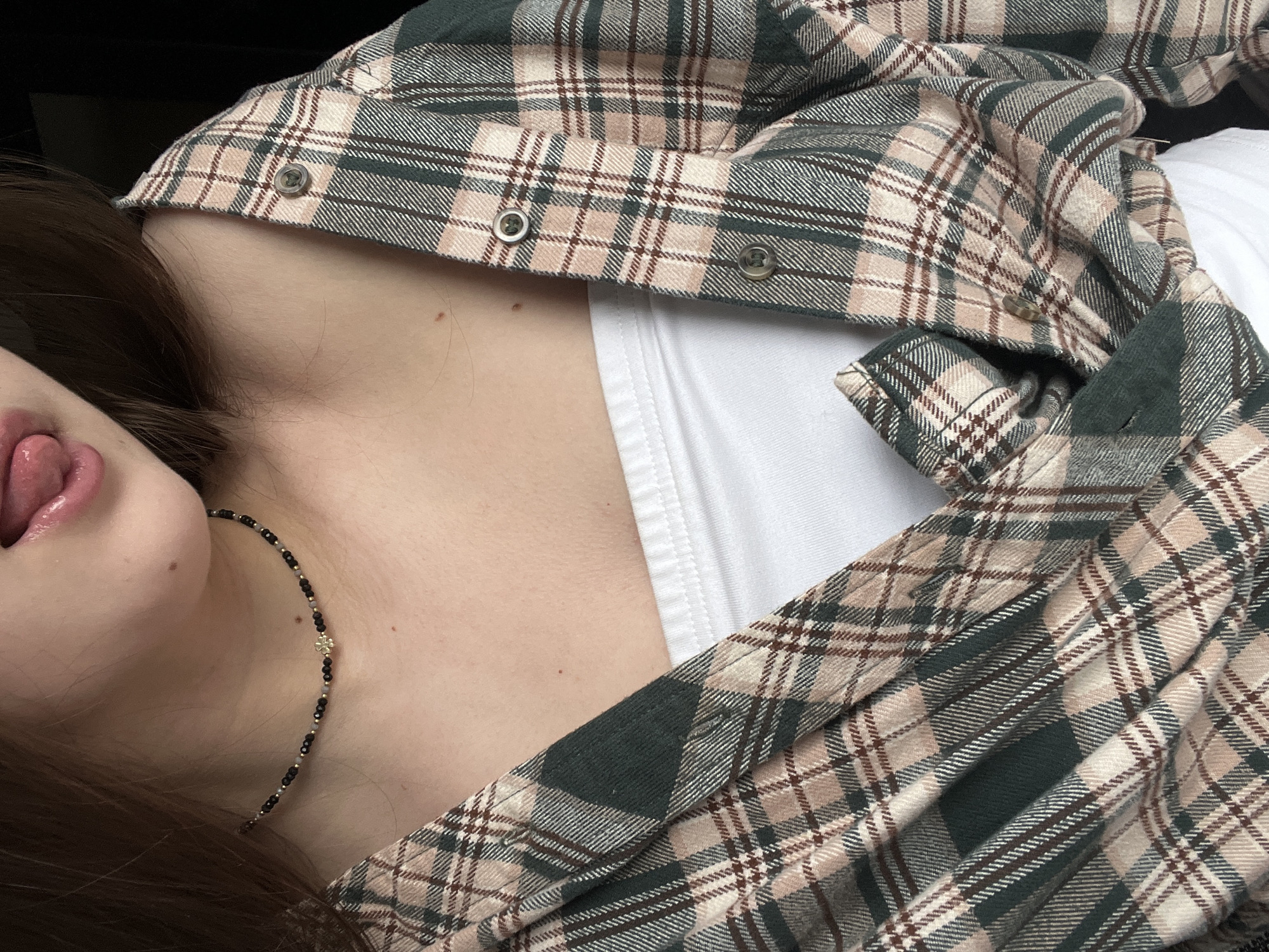 A close-up, angled shot captures the upper torso, neck, and part of the face of a person, who appears to be reclining. The person is wearing a cozy plaid flannel shirt featuring a pattern of dark green, beige, and reddish-brown stripes, worn open over a plain white undershirt or tank top. Several buttons of the flannel shirt are visible along the opening. Around the person's neck is a delicate black and gold beaded necklace, adorned with a small, shiny gold charm that resembles a flower or star. Dark brown hair frames the face, which is partially visible in the lower left, showing the chin, lips, and a playfully extended tongue. The background is dark and out of focus, suggesting an indoor setting. The image conveys a casual and relaxed mood. No text is visible. The time of day, weather, and specific location cannot be determined from the image.