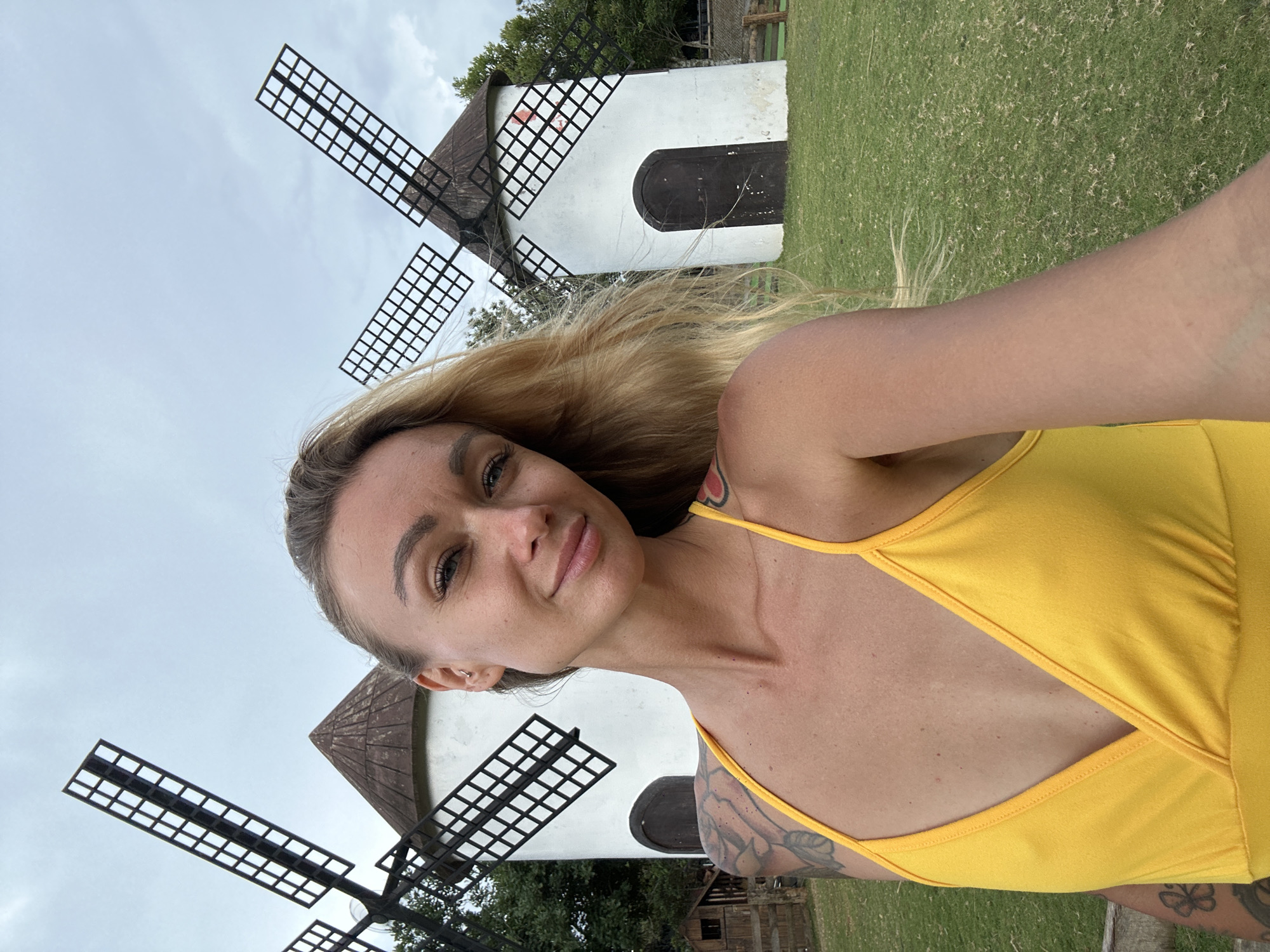 A woman with blonde hair takes a selfie in front of two white windmills with dark, slatted blades. The woman is wearing a bright yellow top with thin straps and a cutout on the chest. She has tattoos on her arm. The windmills are set against a backdrop of green grass and trees, with a cloudy sky above. The image appears to be taken during the day. The scene suggests a rural or tourist location.