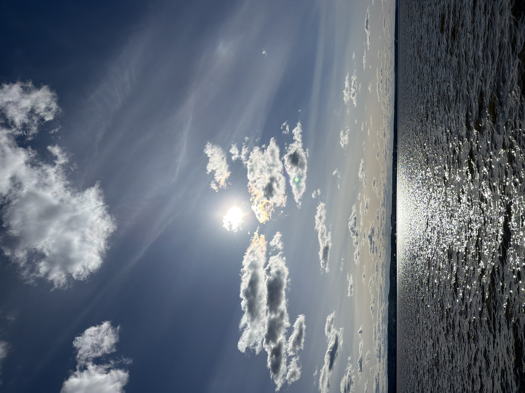 The image shows a sky with clouds and sunlight reflecting on a body of water. The sky is blue with varying cloud formations, some dense and white, others thin and wispy. The sun is partially obscured by clouds, creating a bright glare. The body of water is dark and rippled, with sunlight causing intense reflections. The horizon line is visible, separating the sky from the water. The location is likely near the Votkinsk Reservoir in the city of Votkinsk, Russia, given the expansive body of water and general landscape. No people are visible in the image.