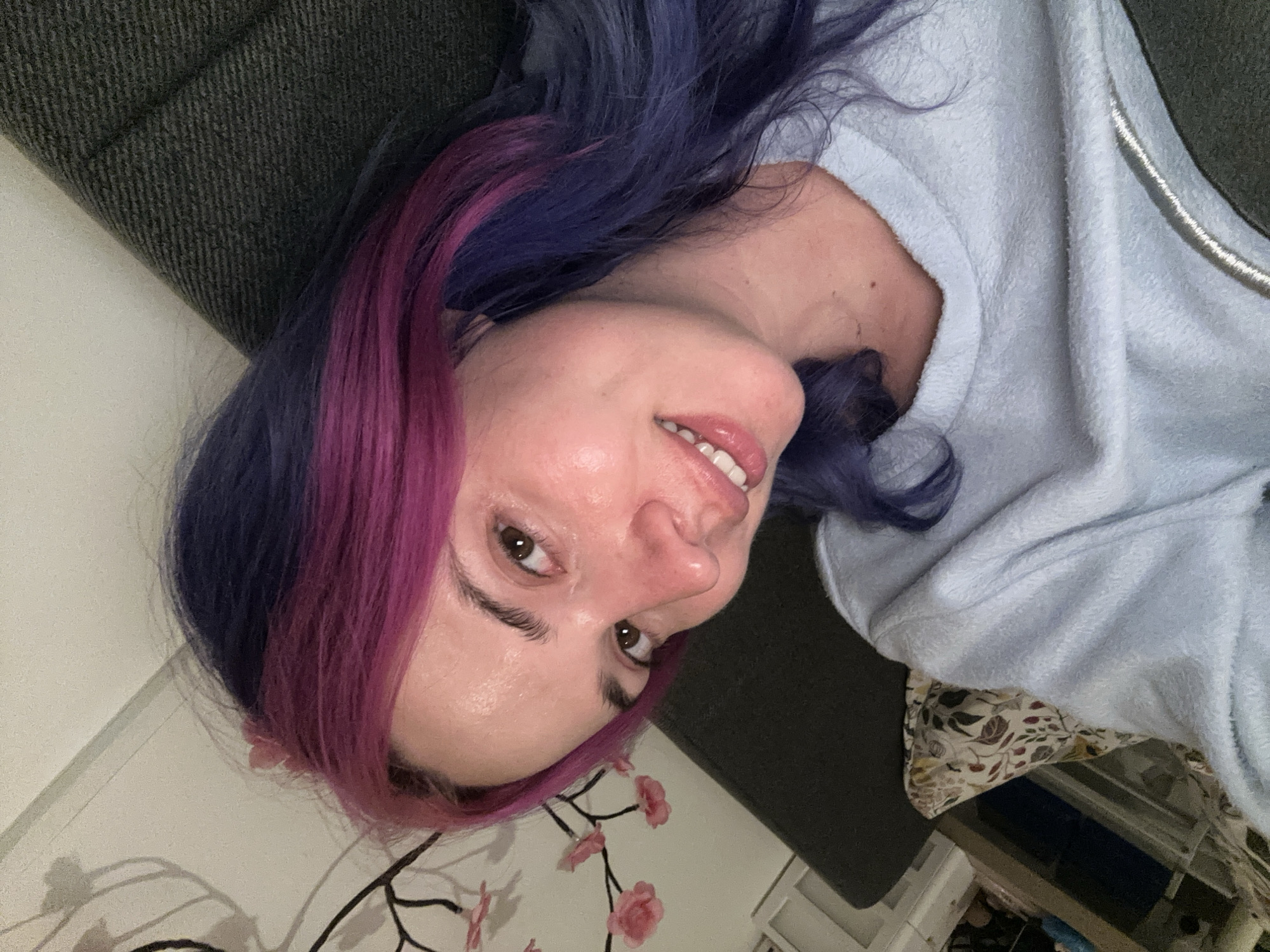 A medium shot, rotated 90 degrees clockwise, features a young woman with long, wavy hair distinctively dyed in purple and magenta/pink streaks. She is lying down, looking directly at the camera with a gentle, inviting smile, revealing her teeth. Her brown eyes are open, complemented by visible eyelashes. She wears a light blue, soft-textured (likely fleece or terry cloth) long-sleeved top, with a silver zipper partially visible on the right side of the image.
The setting is indoors, suggesting a relaxed home environment within the City of Zagreb, Croatia. Behind her head and shoulders is a dark gray, textured fabric, possibly a sofa or cushion. A light-colored wall is visible, adorned in the lower left with a decorative element featuring pink cherry blossoms or similar floral motifs. Underneath her blue top, a section of patterned fabric with a leafy design in cream, brown, and red tones can be seen. The lighting appears to be soft, artificial indoor illumination. The woman conveys a relaxed and friendly demeanor. There is no visible text in the image.