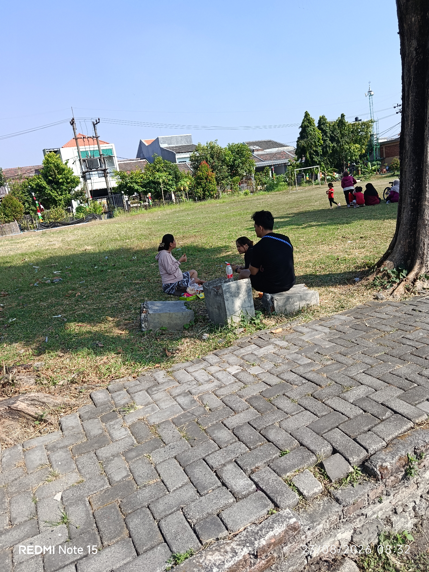 In a grassy field, likely a public park, in Keboansikep, Indonesia, three people are engaged in a picnic. A man and a woman sit on concrete blocks facing each other, with a food container between them. Another person, possibly a child, sits to the woman's right. The surrounding area features a paved walkway, trees, and distant buildings. In the background, several other individuals are visible, some sitting and others standing, enjoying the outdoor space. The sky is clear and blue, suggesting a pleasant day.