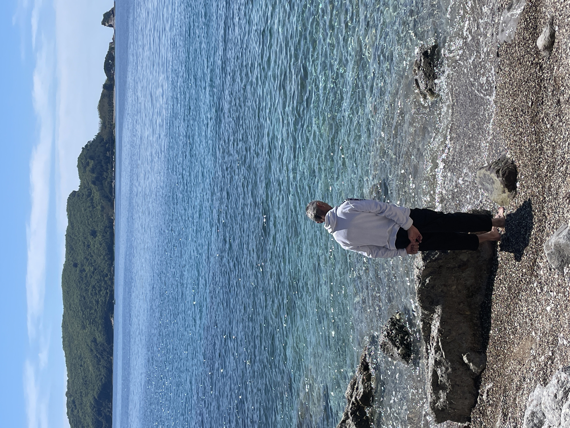 The image shows a person standing at the edge of a beach, looking out at the sea. The person is wearing a white hooded sweatshirt and black pants. The sea is calm and clear, with sunlight reflecting on the surface. A rocky outcrop is visible in the foreground. The location is likely in Budva, Montenegro, given the coastal scenery and rocky terrain, though this cannot be definitively confirmed without more specific details.