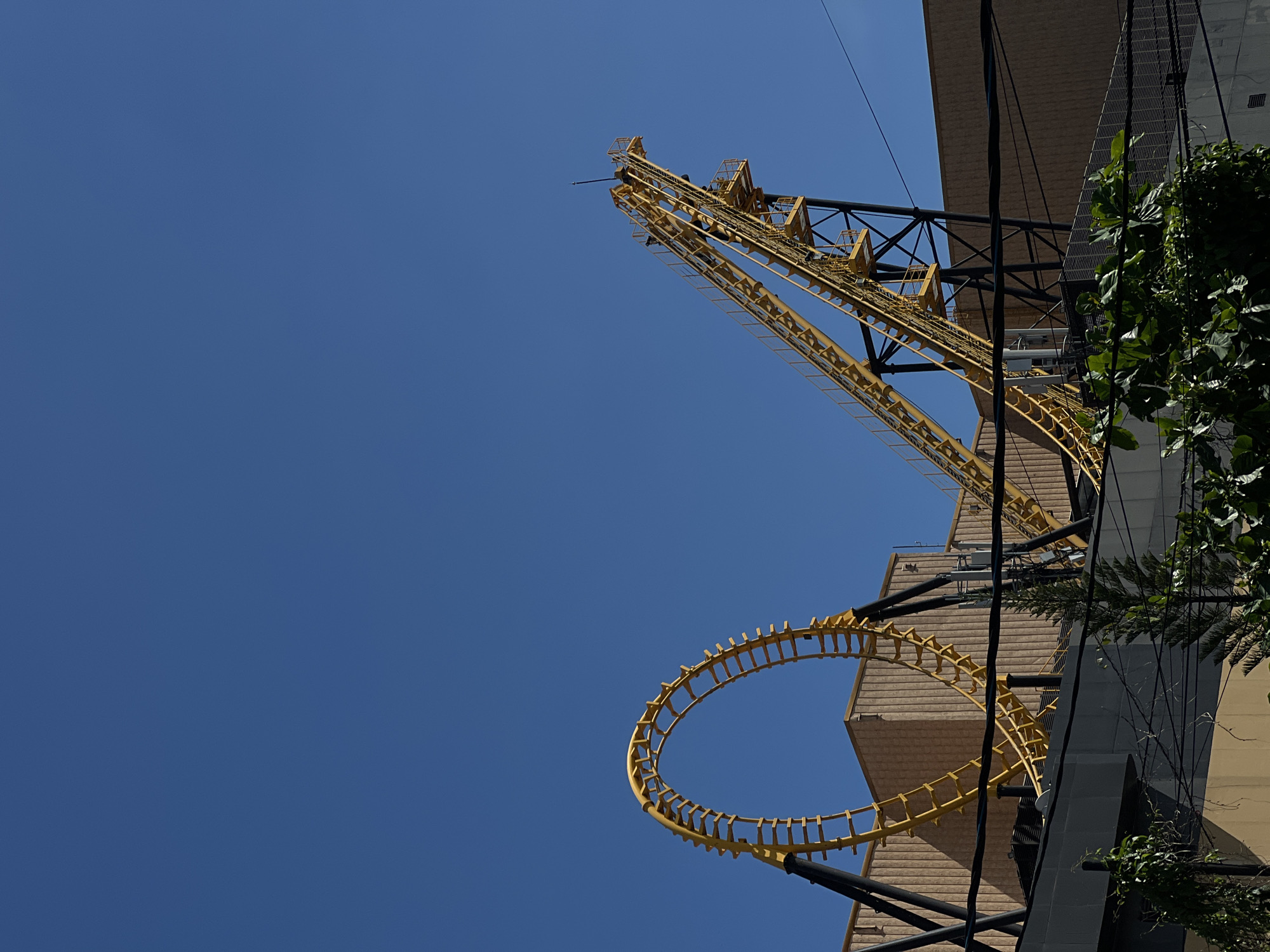 Against a clear, bright blue sky, a striking yellow structure, indicative of a roller coaster or similar amusement ride, dominates the frame. The structure includes an elevated track section, possibly a lift hill, constructed from yellow lattice beams and rungs. Further down and to the left, a distinct circular loop section of the yellow track is prominent.
On the right side of the image, sections of a building are visible, featuring facades with horizontal beige/brown paneling and darker gray sections. Green foliage, possibly from trees or climbing plants, partially covers the right edge of the building, adding a touch of natural contrast. Several dark wires or cables crisscross the scene, passing in front of both the building and the yellow ride structure.
The clear, cloudless sky suggests a sunny daytime setting. There are no people visible in the image, nor is there any discernible text. The scene strongly implies an outdoor amusement park or an entertainment complex.