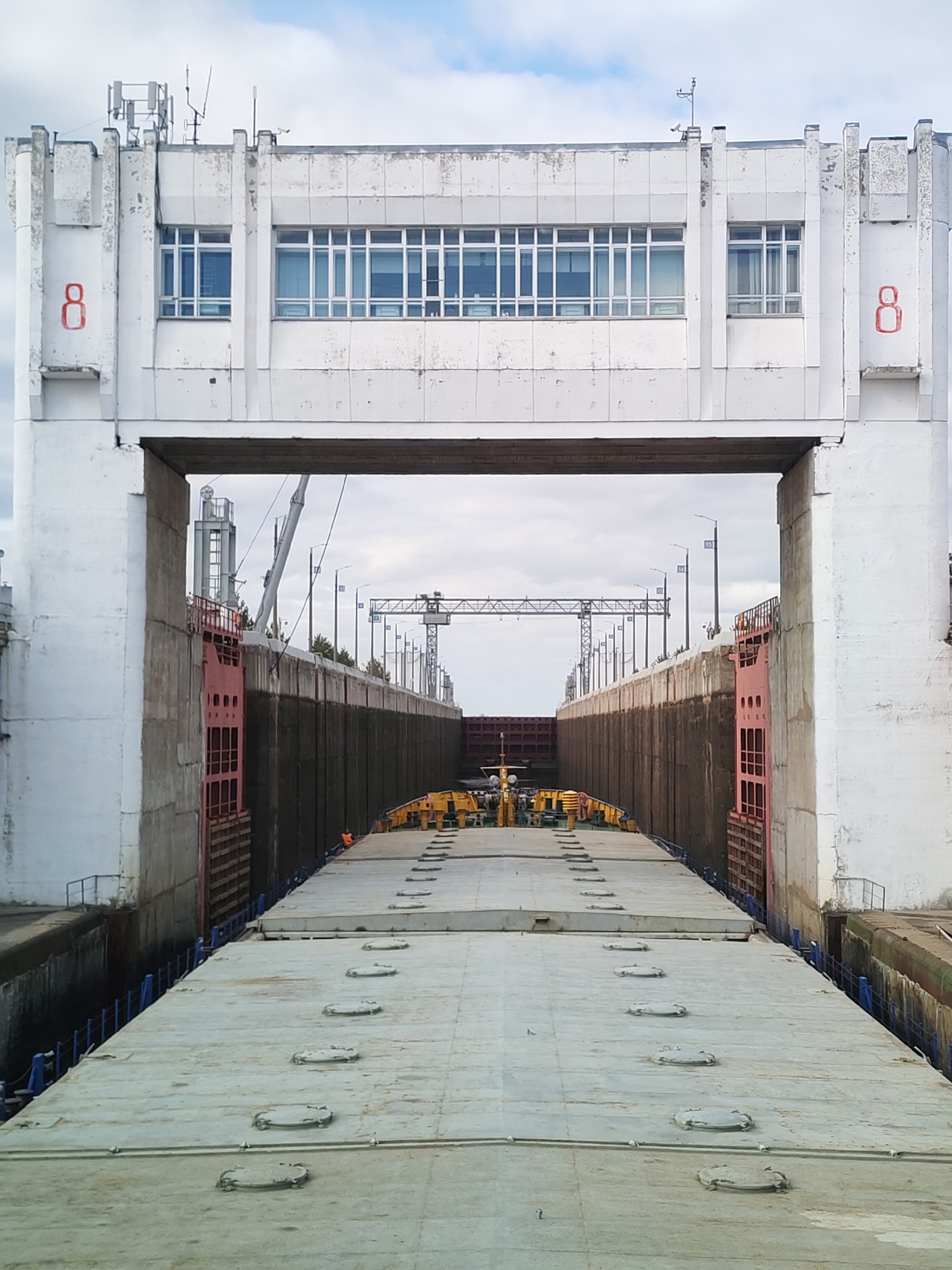 The image depicts the interior of a river lock. The view is looking through the lock structure, with the lock gates and concrete walls enclosing the barge. The lock structure features a white concrete building with windows and antennas on top. There is a large red number 8 painted on both sides of the structure. A massive barge with a grey metal deck fills the lower portion of the image. Along the center of the barge, there are multiple access hatches. At the far end of the lock, a crane is visible. There are also walkways visible on the sides of the lock.