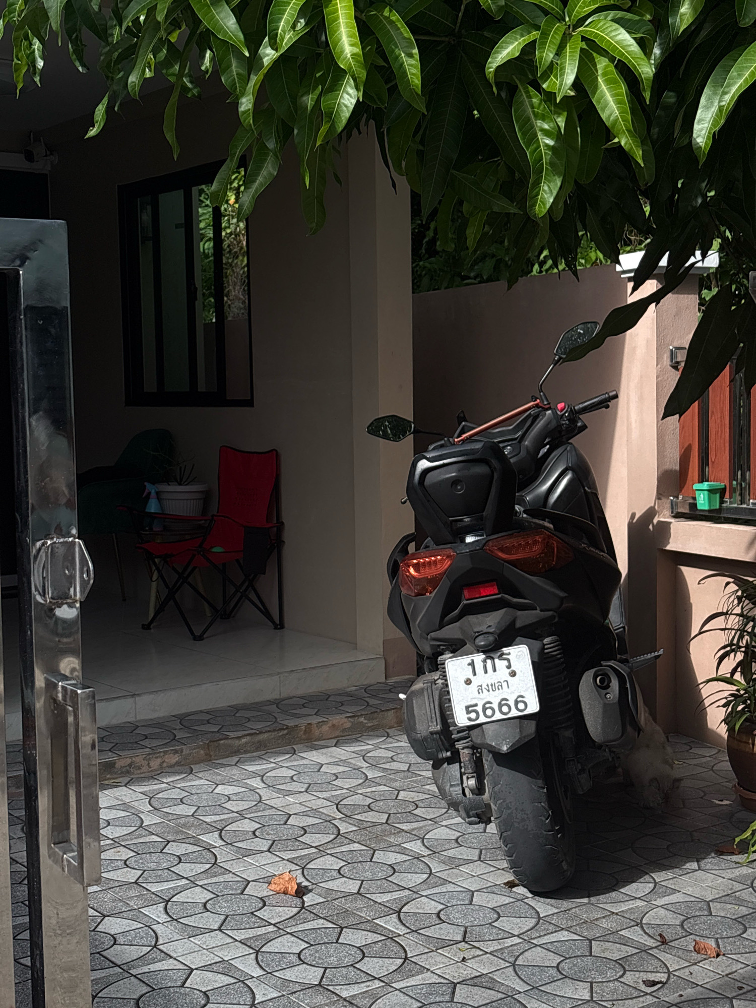 An outdoor scene is depicted, likely in Choeng Thale, Thailand, under bright daylight conditions. A black scooter is parked on a patterned tiled surface in front of a building. The scooter has a license plate with Thai characters and the numbers "1กร" and "5666". To the left of the scooter, a metal gate with a handle is partially visible. Further left, through an open doorway, two folding chairs are present: one is red and appears to be a camping chair, and the other is darker in color and less distinct. Potted plants and foliage from a tree hang overhead, casting shadows on the scene.