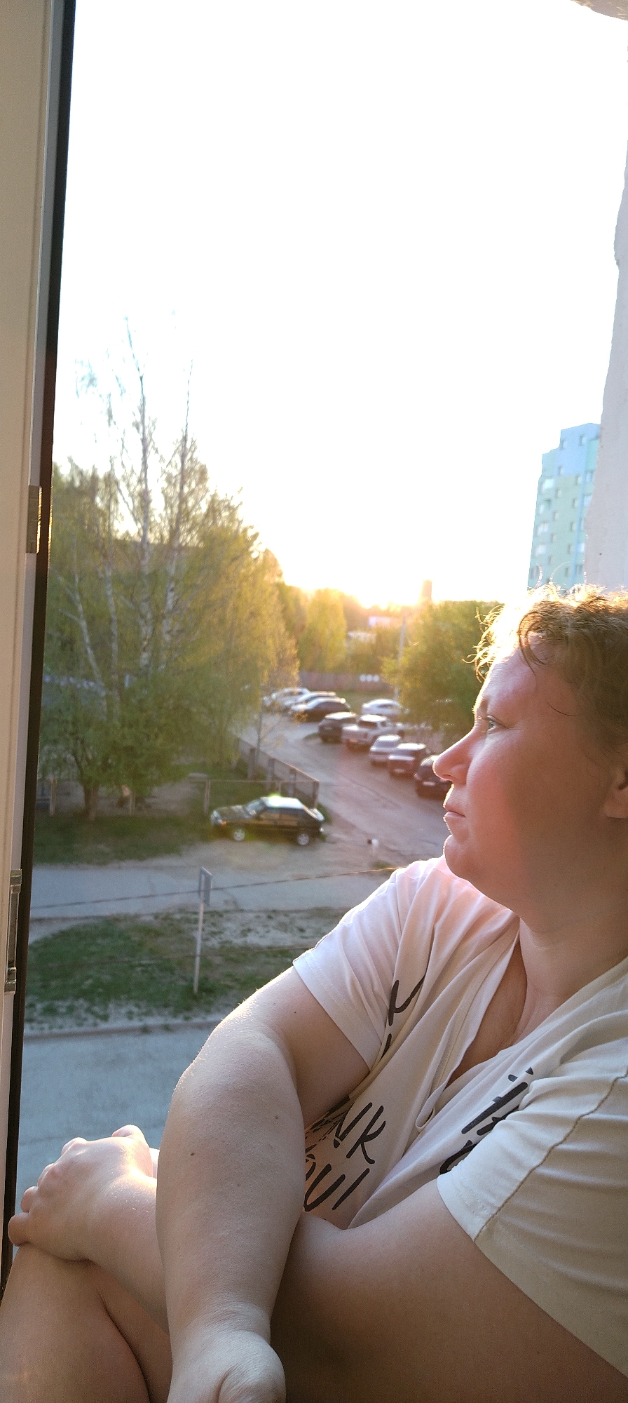 A woman is sitting indoors by an open window, looking out. Outside, there is a view of a street with parked cars, trees, and a building in the distance. The time appears to be sunset. The location is Chaikovsky, Russia.