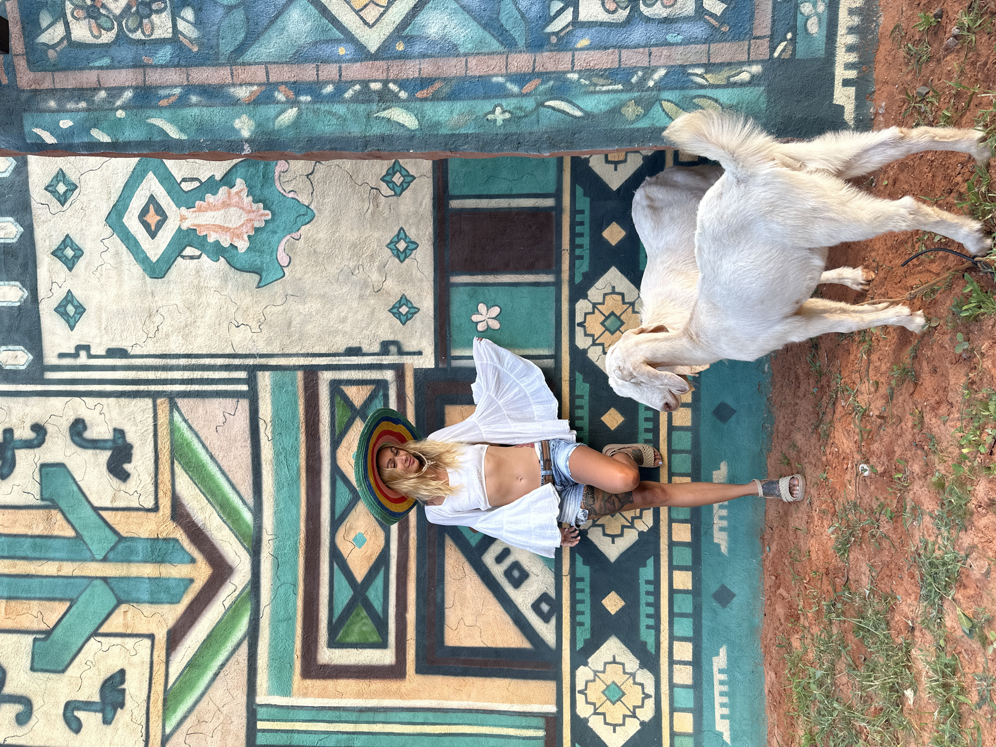 A blonde woman wearing a white crop top, denim shorts, a colorful sombrero, and sandals poses against a vibrantly painted wall with geometric patterns in Mui Ne, Vietnam. Two white goats stand in the reddish-brown dirt in front of her. The wall features a large, intricate mural in shades of teal, peach, brown, and cream, with abstract shapes and tribal-inspired motifs. The woman leans against the wall, her left arm extended, with her head tilted down. One goat is positioned in the foreground, facing away from the camera, while the other is partially visible behind it. The ground is covered with sparse green vegetation.