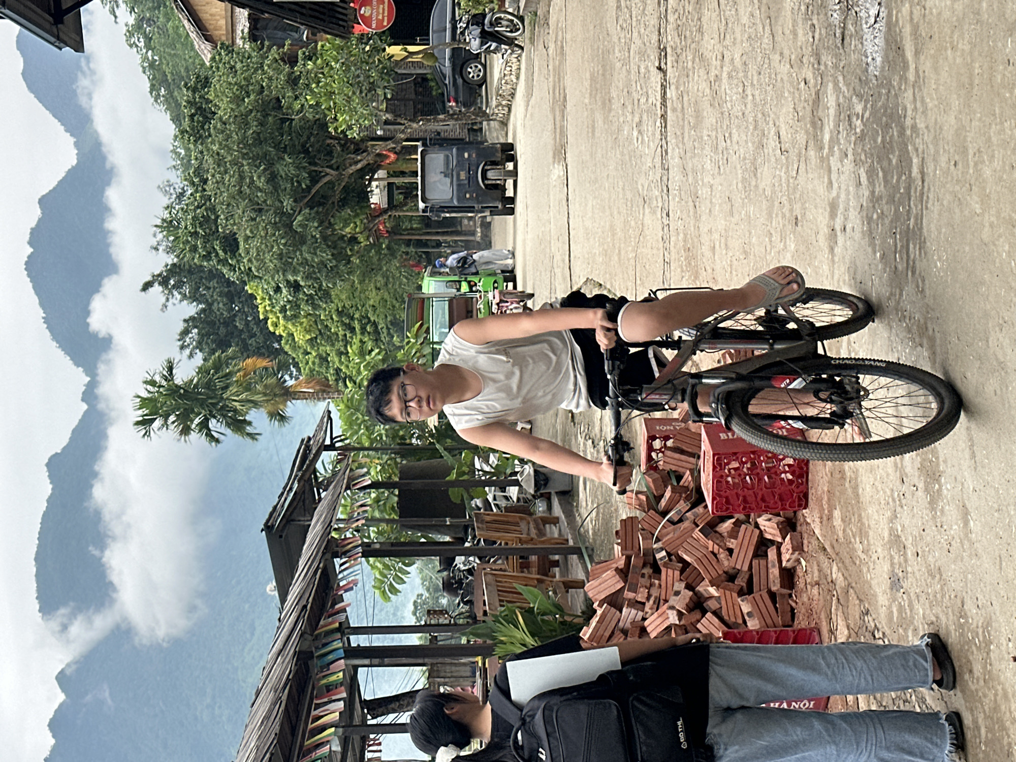 In Mai Chau, Vietnam, a young man wearing a white tank top and black shorts cycles down a dusty, rural road, his right foot in a flip-flop pedaling. Behind him, a young woman with a black backpack on her shoulders, wearing a white and black t-shirt and light blue jeans, stands facing away from the camera. To their left, a large pile of red bricks and red crates are stacked on the side of the road. In the background, a traditional wooden building with a thatched roof, adorned with colorful flags, stands near lush green trees and the misty peaks of mountains characteristic of the region. Several vehicles, including a black tuk-tuk and a green passenger van, are visible further down the road, suggesting a bustling but unpaved commercial or residential area. The sky is overcast, with clouds partially obscuring the mountain range.
