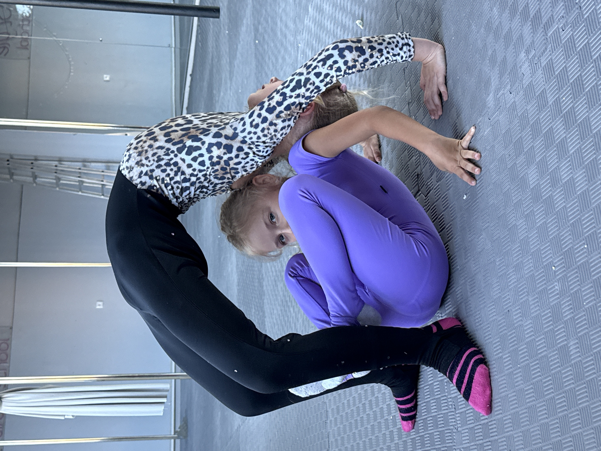 Two young girls are practicing gymnastics on a grey, textured mat floor. The girl on the left is wearing a leopard-print long-sleeved leotard and black leggings. She is bent backward in a deep bridge, with her hands on the floor. The girl on the right is wearing a purple long-sleeved leotard and black socks with pink stripes. She is in a crouched position, facing the other girl. Her head is tilted and she is looking towards the camera.
In the background, there are metal poles and what appears to be part of a rack or stand with folded white material. The setting seems to be an indoor space, possibly a gymnastics studio or a similar training facility. The lighting is bright, suggesting it is daytime. No specific weather cues are visible. The overall mood appears to be one of concentration and athletic activity. There is no visible text in the image.