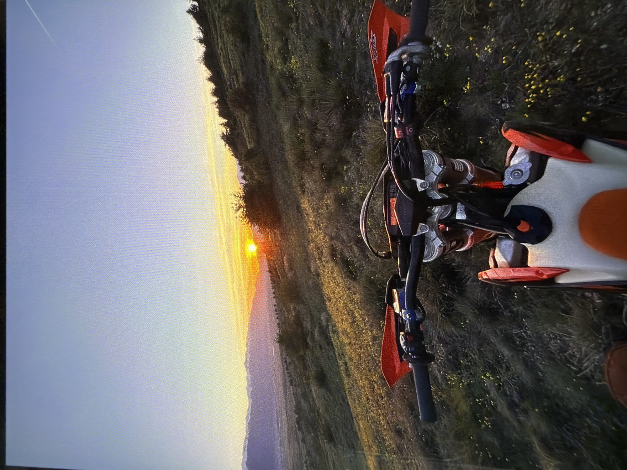 This media file captures a sunset over a mountainous landscape, with a dirt bike prominently featured in the foreground. The image is taken from a slightly elevated perspective, looking down and across the scene.
The main subject is an orange and white dirt bike, positioned on the right side of the frame, angled towards the left. The handlebars, front forks, parts of the engine, the white seat, and the orange rear fender are clearly visible. The handguards are orange, and the brand "KTM" is discernible on one of them. The "WP" logo, indicating WP Suspension components, can be seen on parts of the front fork assembly. The bike is parked on uneven, grassy terrain dotted with small yellow flowers.
In the background, a vast panorama unfolds, dominated by a setting sun on the horizon. The sun's intense orange glow illuminates the sky and casts a warm hue over the distant mountains and valleys. The sky transitions from a bright orange near the sun to softer yellows and blues higher up. A faint white contrail from an aircraft is visible in the upper left portion of the sky. The landscape consists of rolling hills and mountains, covered in dark green vegetation, likely forests or fields. The scene suggests a rural or wilderness setting, fitting the provided location context of Razlog, Bulgaria, which is surrounded by mountain ranges like Pirin and Rhodope.
The time of day is clearly sunset, indicated by the sun's low position and the long shadows, creating a serene and picturesque atmosphere. The weather appears clear and calm. The dirt bike is stationary, suggesting a pause during an off-road ride to enjoy the scenic view, or the end of a day's adventure. The image exhibits visible horizontal scanning lines, indicating it was likely a photograph taken of a digital screen or monitor.
