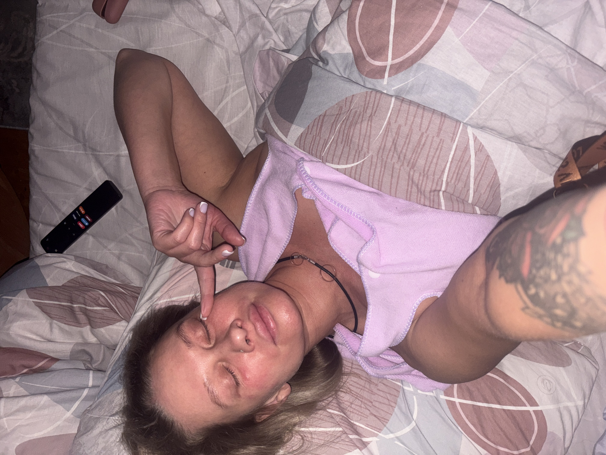 A fair-skinned woman lies on a bed with patterned bedding, touching her eyelid with her right index finger. She wears a light purple top and a black choker. A remote control is visible on the bed beside her. The arm closest to the viewer has a tattoo. The location is likely in Perm, Russia.