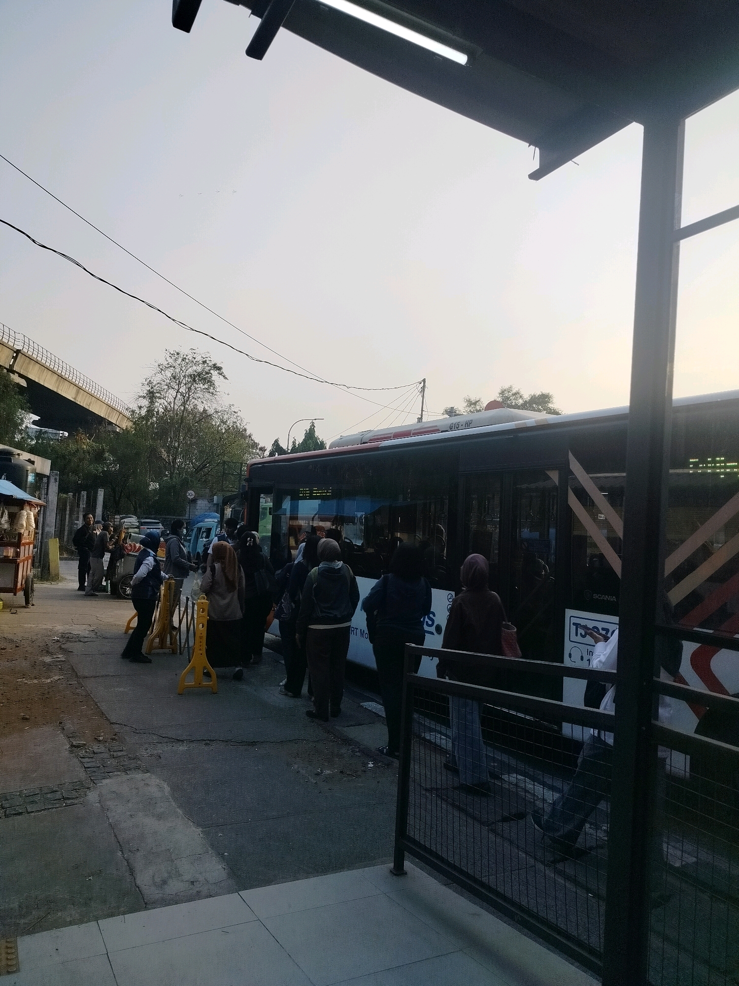 A group of people are waiting in line for a bus at a bus stop in East Jakarta, Indonesia. The bus is a red and white Scania model. The sky is overcast, and there are power lines visible in the background. There is a food cart to the left of the bus stop, and a highway overpass in the distance. The people in line are dressed in casual clothing, and most of them are wearing masks. The bus stop has a metal fence and a covered shelter.