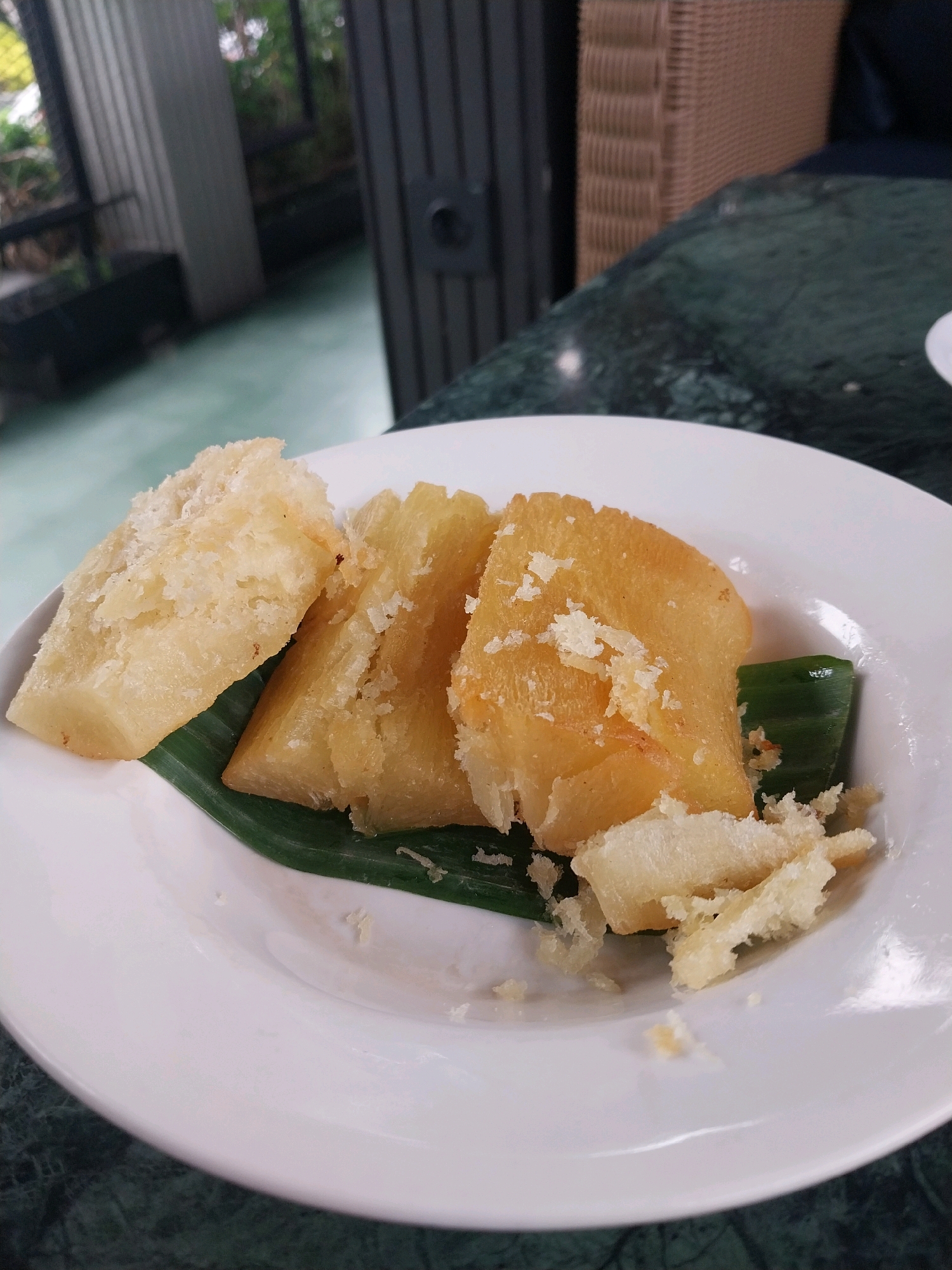 A close-up shot shows three triangular pieces of fried cassava, sprinkled with grated coconut, arranged on a white plate lined with a green banana leaf. The cassava pieces are golden brown and appear crispy. The plate sits on a dark green marble table. In the blurred background, a dark-colored rattan chair and outdoor greenery are visible, suggesting the scene is set at an outdoor dining area. The setting appears to be a casual eatery or cafe in Bugisan, Indonesia.