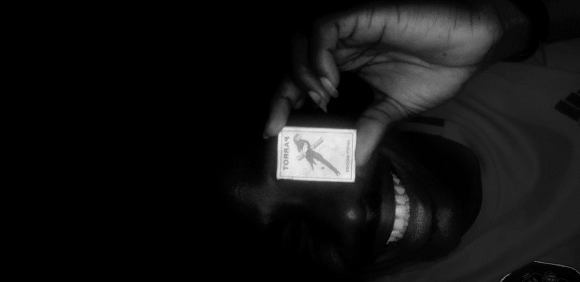 The image shows a dark-skinned person holding a matchbox in front of their face. The matchbox has the word "PARROT" printed on it. The person is smiling, revealing their teeth. The background is dark and out of focus, but hints of clothing suggest they may be wearing a t-shirt. No information can be gathered from the image regarding location in Narayi, Nigeria.