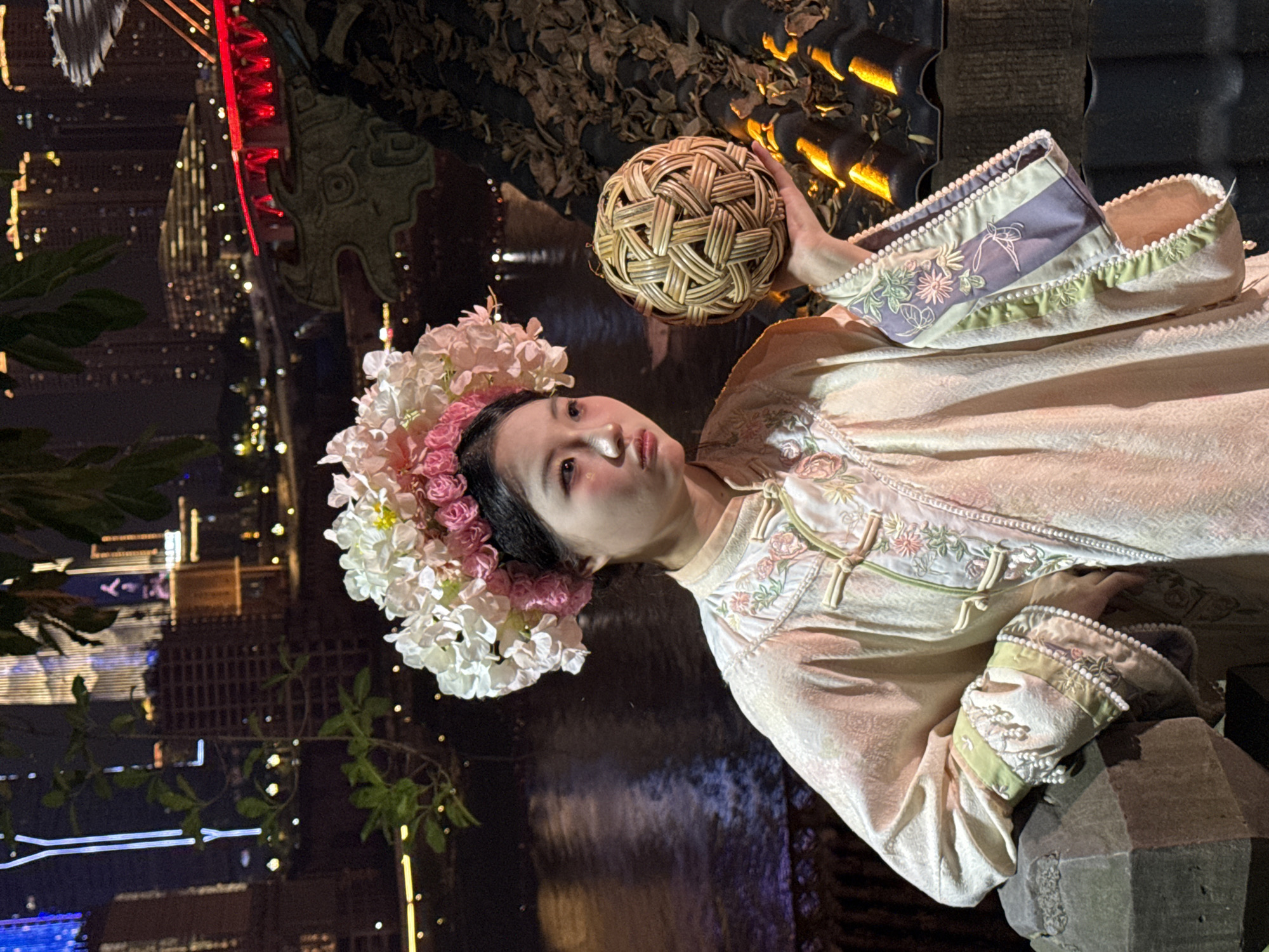 A woman wearing traditional Chinese clothing and a floral headdress is posing with a woven ball in Yuzhong District, China. The location appears to be outdoors at night, with blurred city lights and reflections in the background. The woman is positioned slightly off-center, looking directly at the camera. Her outfit is embellished with floral patterns and decorative trim. In the upper background is a rock structure with lights.