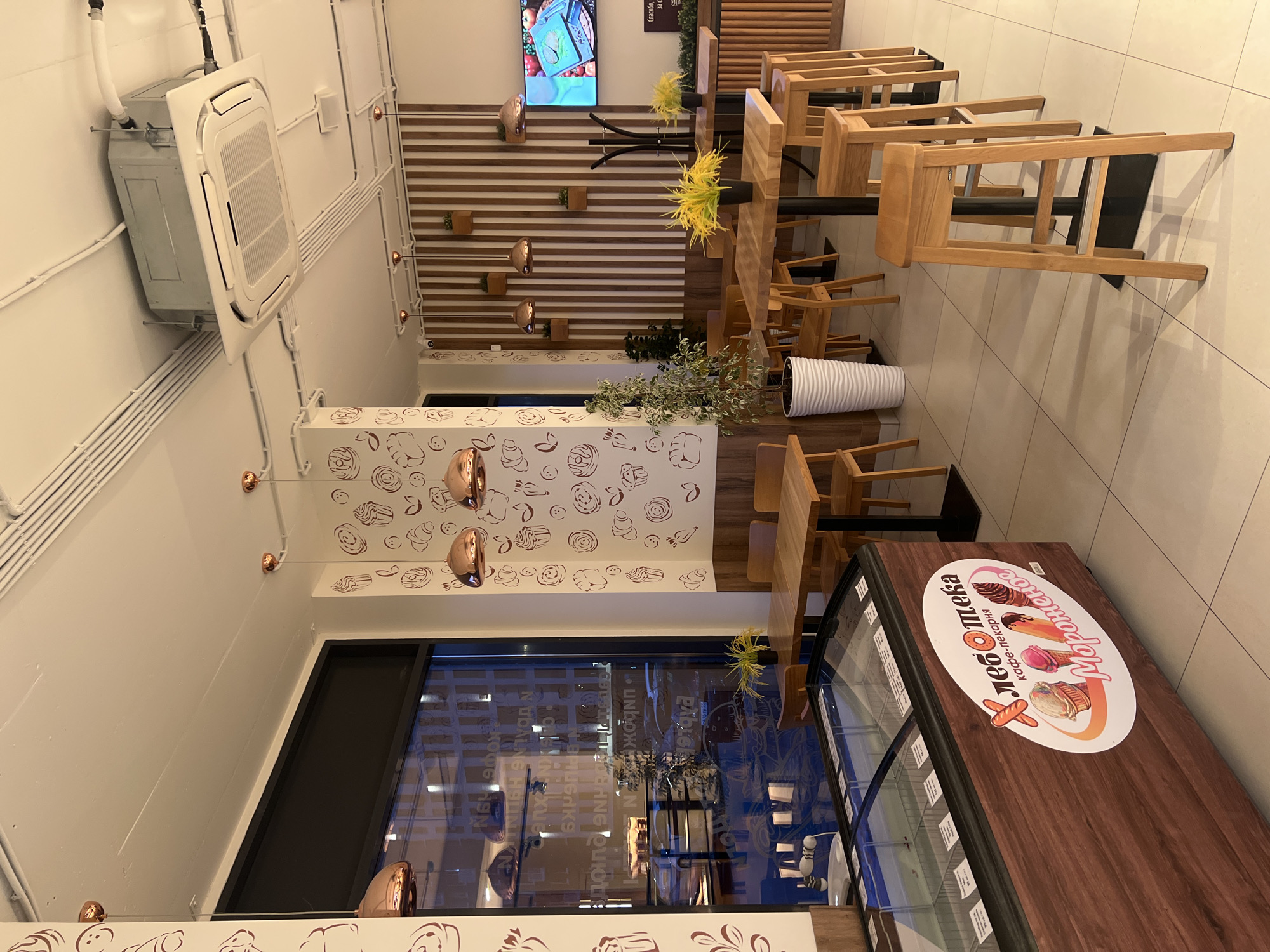 The image captures the interior of the "Hleb Oleka" cafe-bakery in Tyumen, Russia. The view is from a slightly elevated angle, looking down the length of the establishment.
On the right is a wooden-topped display case with "Hleb Oleka" branding. Tables with light wooden chairs are arranged in rows, leading towards a back wall with horizontal wooden slats and decorative sconces. Above the slats, there's a screen showing what appears to be a food-related video. A white column with decorative donut and pastry prints is on the left. Large windows offer an ambiguous view of a city landscape with lights at night. An air conditioning unit is installed on the ceiling. The scene is brightly lit.