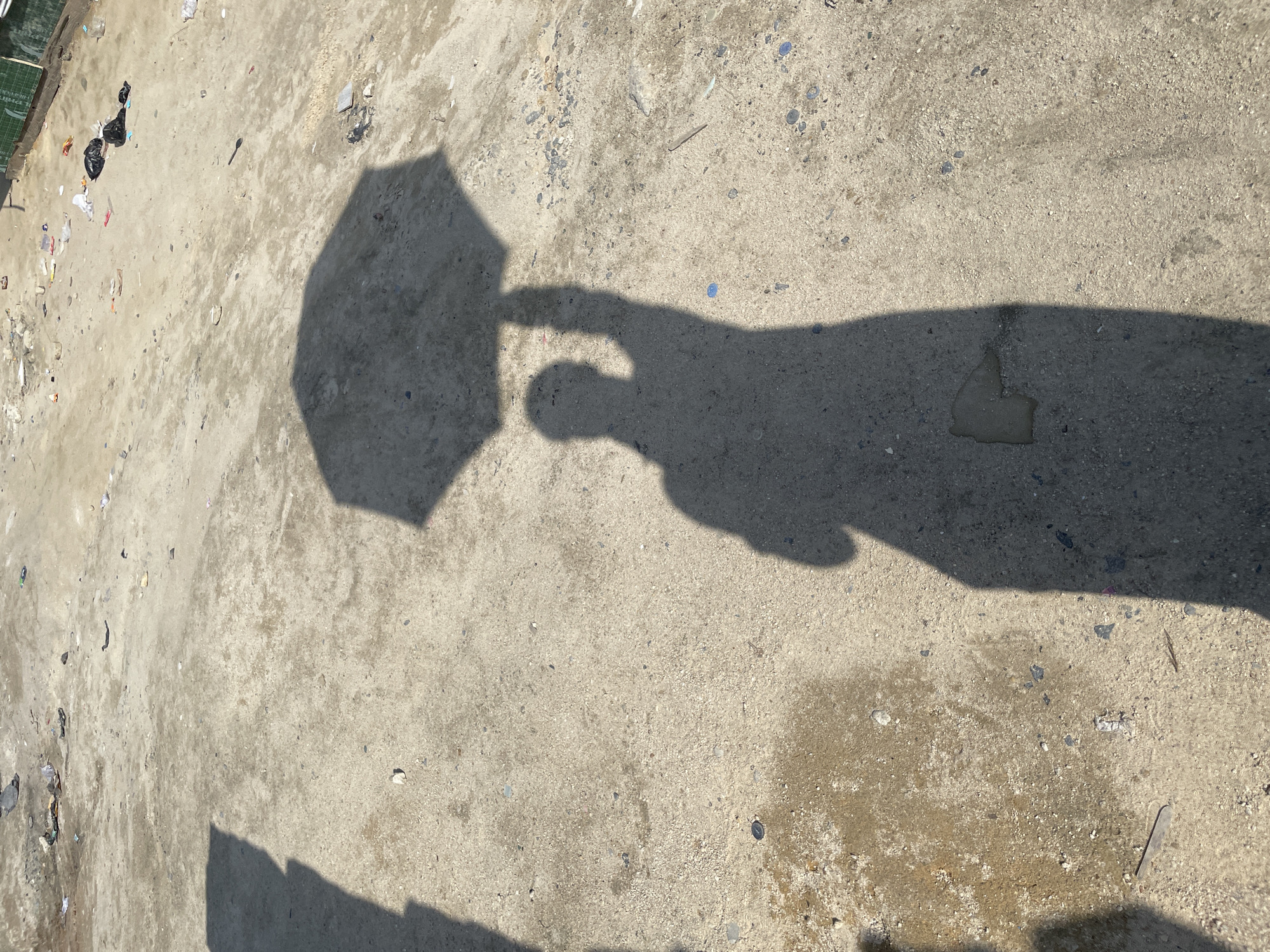 In Iranla, Nigeria, a shadow of a person holding an umbrella is cast on a dirty, paved area. The ground is covered in debris, including plastic bags, wrappers, and other litter. The shadow of the person is elongated and distorted by the low angle of the sun, obscuring details of their features. The umbrella's shadow is clearly visible above the person's head. The environment appears to be an outdoor space, possibly a street or a vacant lot, that is not well-maintained.