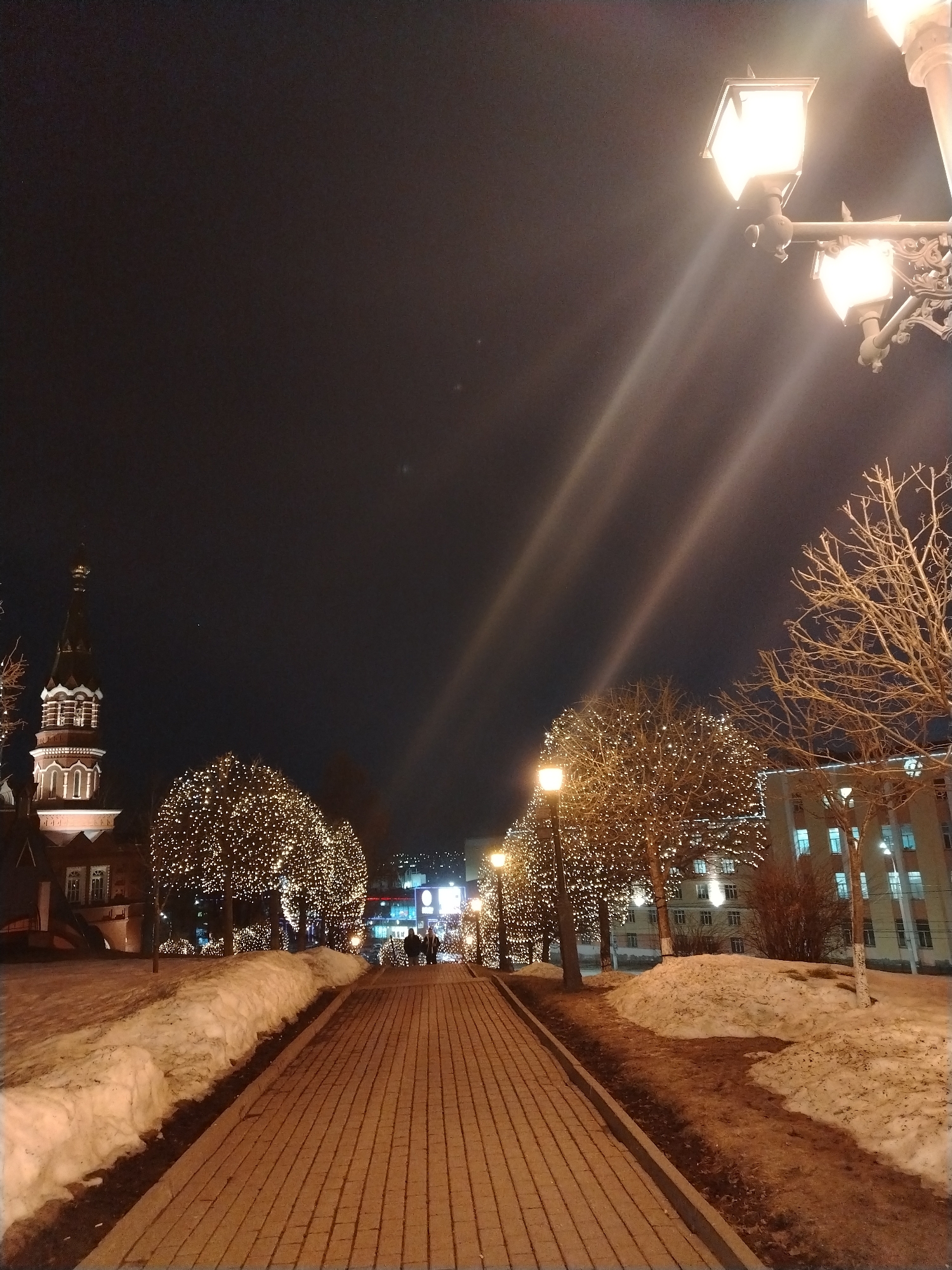 Captured at night in Izhevsk, Russia, the image features a brick-paved pedestrian walkway lined with snow-covered banks on either side. Trees illuminated with strings of lights flank the walkway, leading towards figures in the distance. To the left, a church with a distinct spire is visible, while a building with lit windows appears on the right. Street lamps cast light beams upward, illuminating the dark sky. A decorative lamppost is prominent in the foreground.