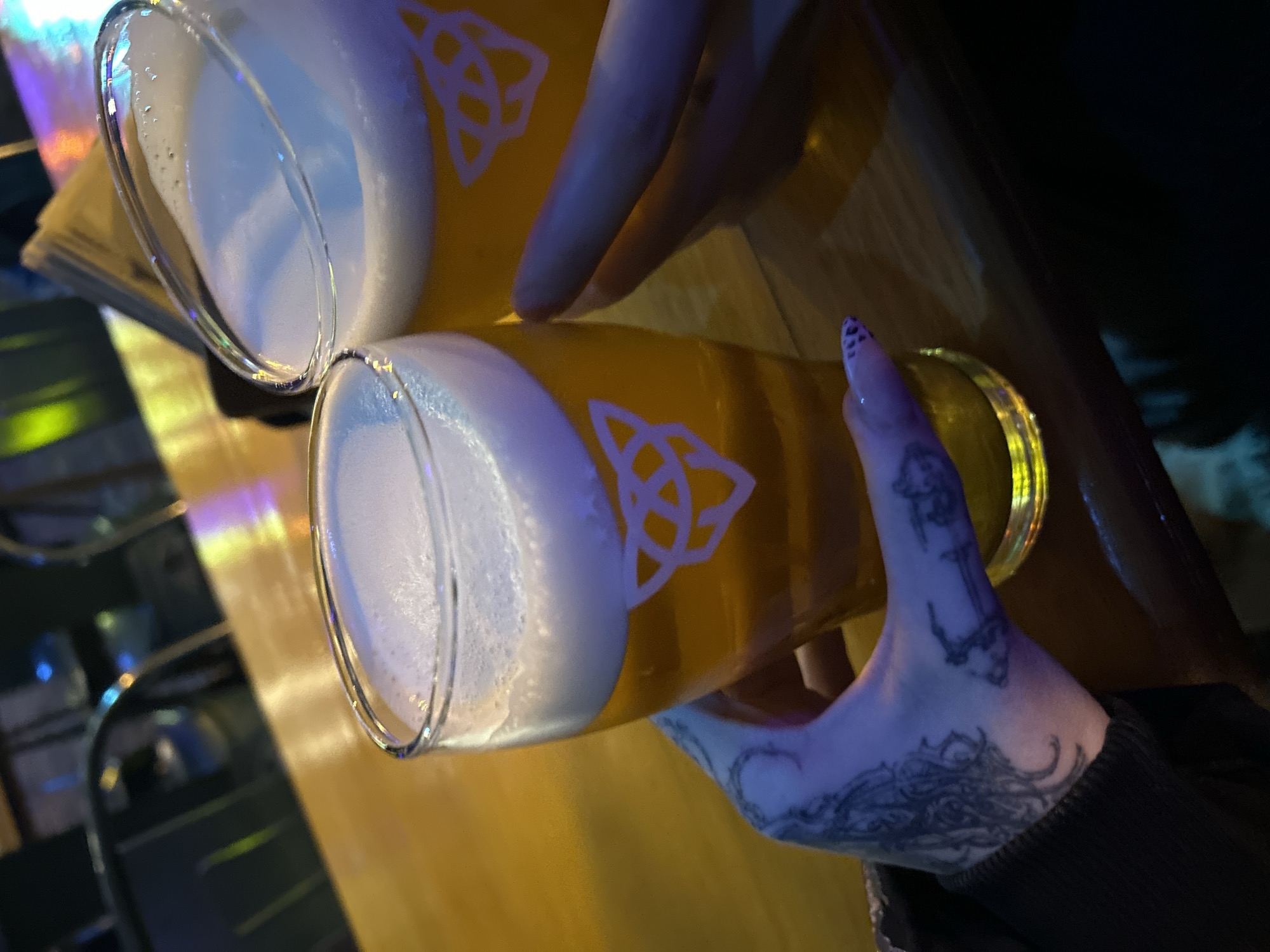 The image displays a close-up, high-angle shot of two hands holding glasses of beer, likely in a dimly lit bar or pub environment. The glasses are made of clear glass, filled with a golden-amber beverage, and topped with a substantial, frothy white head. Each glass features a distinct white Celtic knot design on its side.
On the right side of the frame, a hand with extensive and intricate black tattoos covering the back of the hand and extending onto the wrist firmly grasps one of the beer glasses. The fingernail on the visible finger has a dark, patterned polish. Another hand is partially visible in the upper left, holding the second glass.
The background is dark and out of focus, showcasing blurred reflections of colorful artificial lights, including hues of purple, yellow, and blue, hinting at a lively atmosphere. The surface on which the glasses are presented appears to be a glossy table or bar top, reflecting the ambient light. The scene suggests an evening or nighttime gathering. No discernible text is visible other than the Celtic knot symbols on the glassware.