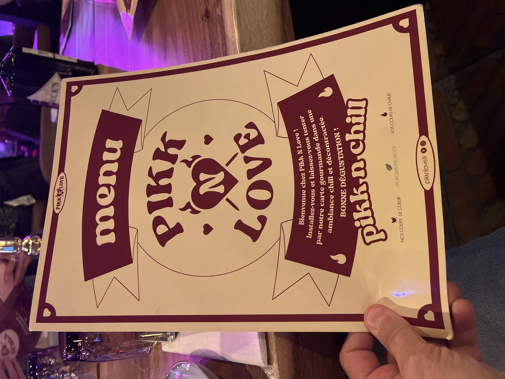 The image shows a menu from "Pikk N Love", a restaurant in Saint-Jean-de-Védas, France. The menu is held by a hand, displaying a beige background with a maroon border. The text reads "menu Pikk N Love pikknchill". The menu also includes a welcome message in French, indicating a chill and relaxed atmosphere with tasty food. The menu further shows the restaurant's website and social media handles.
