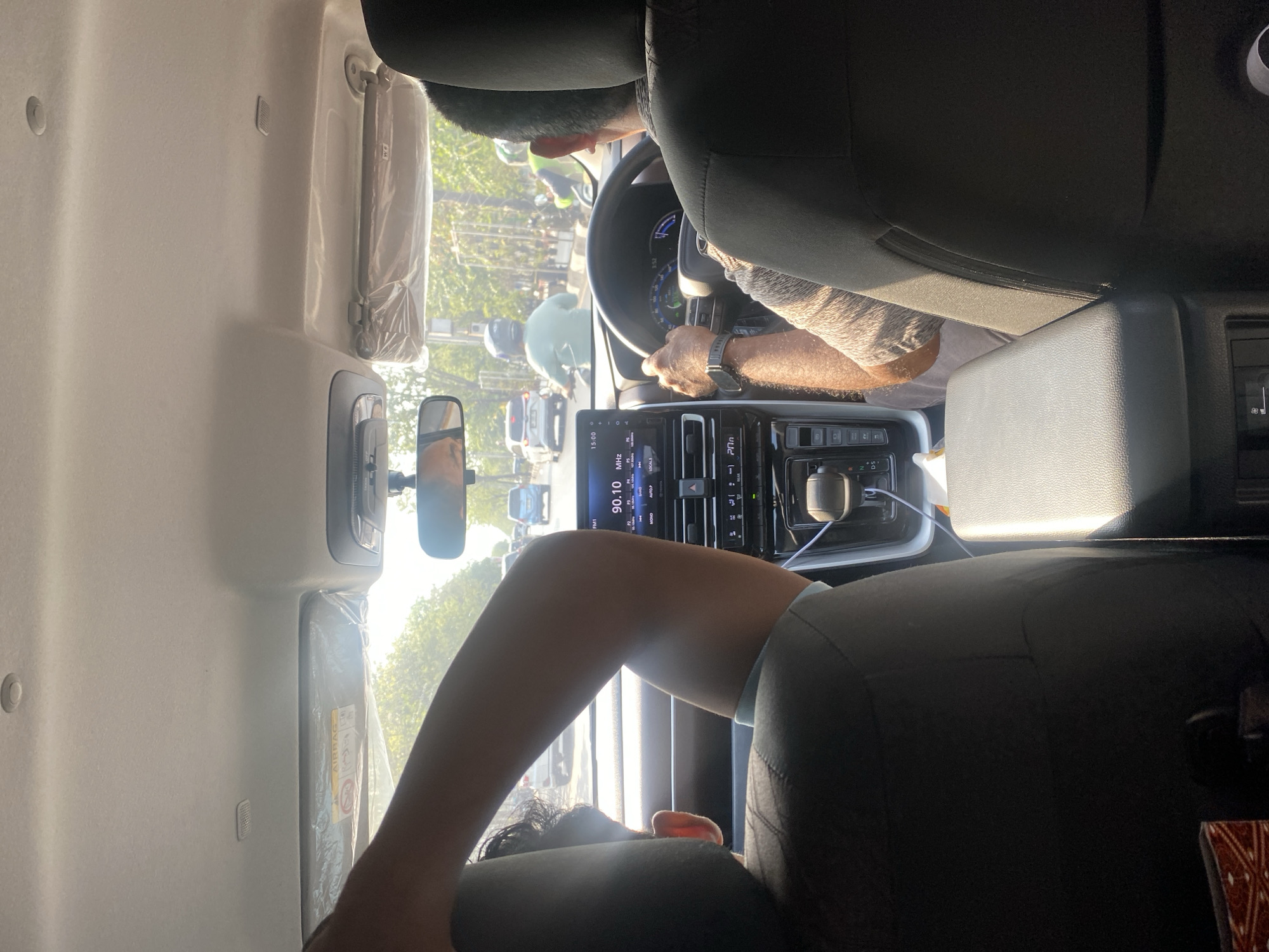 The image captures the interior of a car, seen from the back seats, as it travels through the city of Banjarmasin, Indonesia. Sunlight streams in through the windows, illuminating the dashboard and the driver. The driver, a man with visible arm hair, is holding the steering wheel. To the left, another person's arm is visible, resting on the back of their seat. The car is in traffic, with other vehicles, including cars and motorcycles, visible on the road ahead. The dashboard features a digital display showing "90.10 FM" and other information, along with a gear shift and various buttons. Trees and buildings line the street outside, suggesting an urban environment.