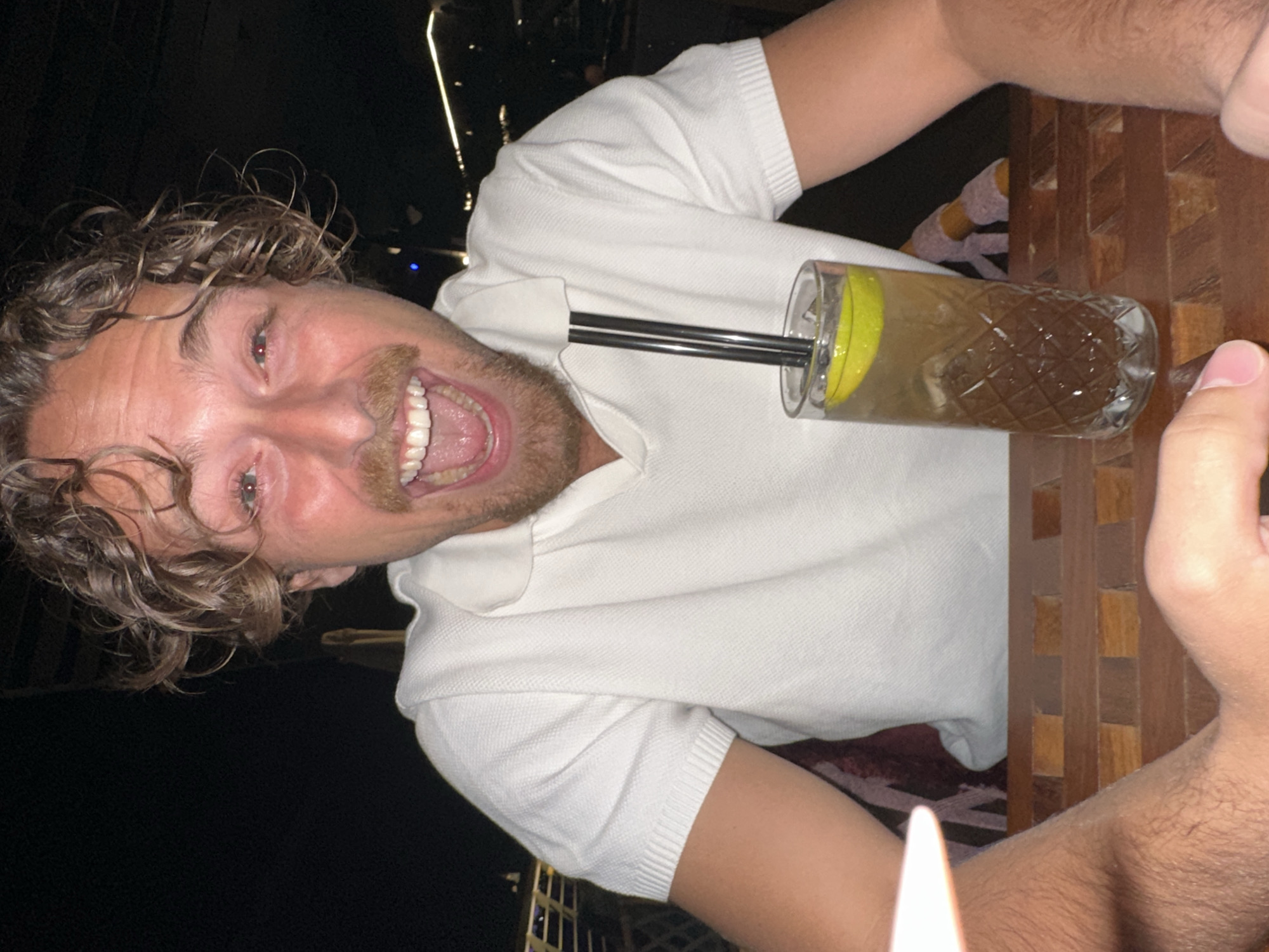 A man is sitting at a wooden table outdoors at night. He has curly brown hair, a mustache, and is wearing a white polo shirt. He is smiling with his mouth open and holding a drink with two straws and a lime slice. In the background, there are blurred lights and structures, possibly indicating a restaurant or bar in Belgrade, Serbia.