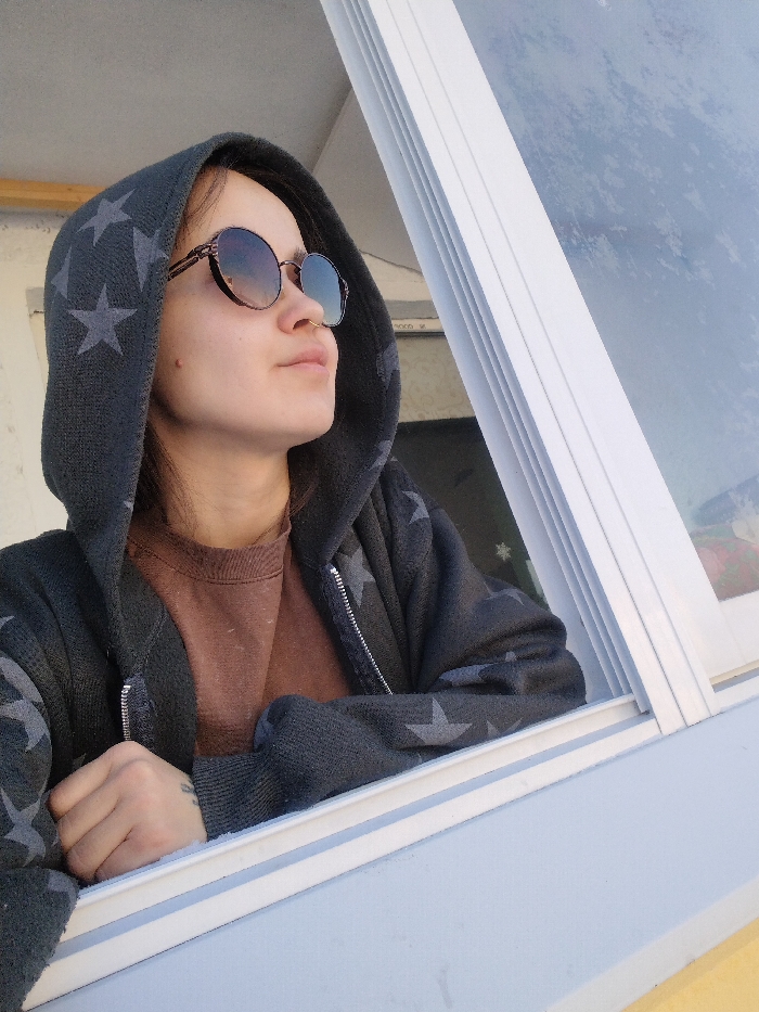 An individual, appearing female, is seen in a medium shot, looking upwards and to the right through a partially open window. She wears a dark gray hooded sweatshirt patterned with lighter gray stars and a partially visible zipper, over a plain brown long-sleeved shirt. Round, gradient-tinted sunglasses cover her eyes, and a small metallic ring is present on her left nostril. A small, dark mark is visible on her right cheek. Her left forearm rests on the light blue window frame, with a tattoo visible on the inner wrist. The window consists of white frames; the right pane displays a diffuse white and light blue reflection or frost, while the open section to the left reveals a plain, light-colored exterior wall. The interior wall visible on the left is a light, off-white hue. The scene takes place in Noviy, Russia.