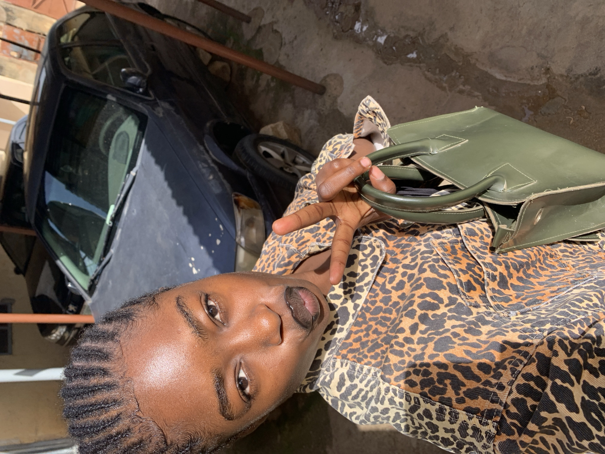The image shows a person in a leopard print jacket, holding a green purse. The person is making a peace sign with their fingers. They are in an outdoor setting, with a car visible in the background. The ground appears to be dirt and some wet patches, suggesting it might have rained recently. The lighting suggests it is daytime. The person's face is partially visible, and they have dark skin and braided hair. The overall mood of the image is casual and self-expressive. There is no visible text in the image.