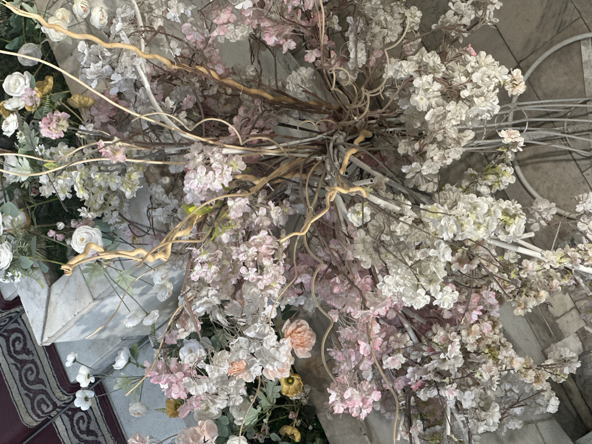 A close-up, top-down view of a large floral arrangement featuring many delicate artificial flowers in shades of white, pink, and cream. The flowers are clustered together on thin, brown, twig-like branches. Some of these branches appear to be real, while others are likely artificial and painted white. The arrangement is dense, with flowers and branches obscuring much of the background. In the upper left corner, a portion of a patterned red rug is visible, along with a light-colored stone structure. The overall impression is one of a decorative display, possibly for an event or as interior decoration. The location is not identifiable from the image itself and is stated to be in Bishkek City, Kyrgyzstan.