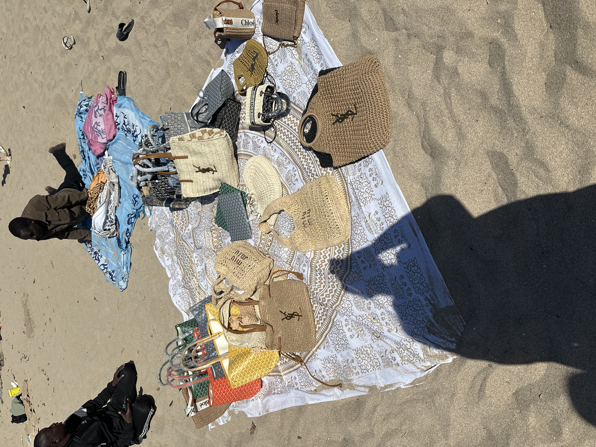 The image shows a vendor displaying handbags on a beach in Sant Josep de sa Talaia, Spain. The scene includes various straw and designer-style handbags, some marked with "Chloe" and "YSL" logos, arranged on a white patterned cloth spread on the sand. A person in dark clothing sits on the left side of the frame, appearing to be observing or guarding the merchandise. Discarded flip-flops are scattered on the sand nearby. The environment consists of sandy beach terrain and direct sunlight.
