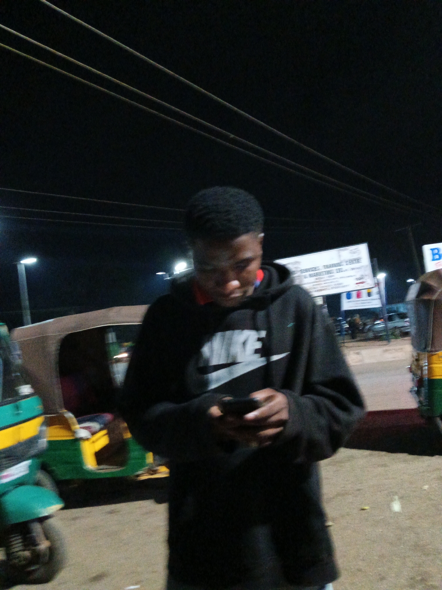 The image captures a person in a black Nike hoodie, looking down at their phone, standing outdoors at night. The setting appears to be a street in Bukuru, Nigeria, indicated by the presence of auto-rickshaws, commonly known as "keke" or "marwa" in Nigeria, and overhead power lines. Streetlights illuminate the scene, casting a dim glow. In the background, a sign for "SERVICES, TRAINING CENTRE & MARKETING LTD" is visible. The ground is dirt or gravel, suggesting an unpaved area. The overall atmosphere is casual and nocturnal.