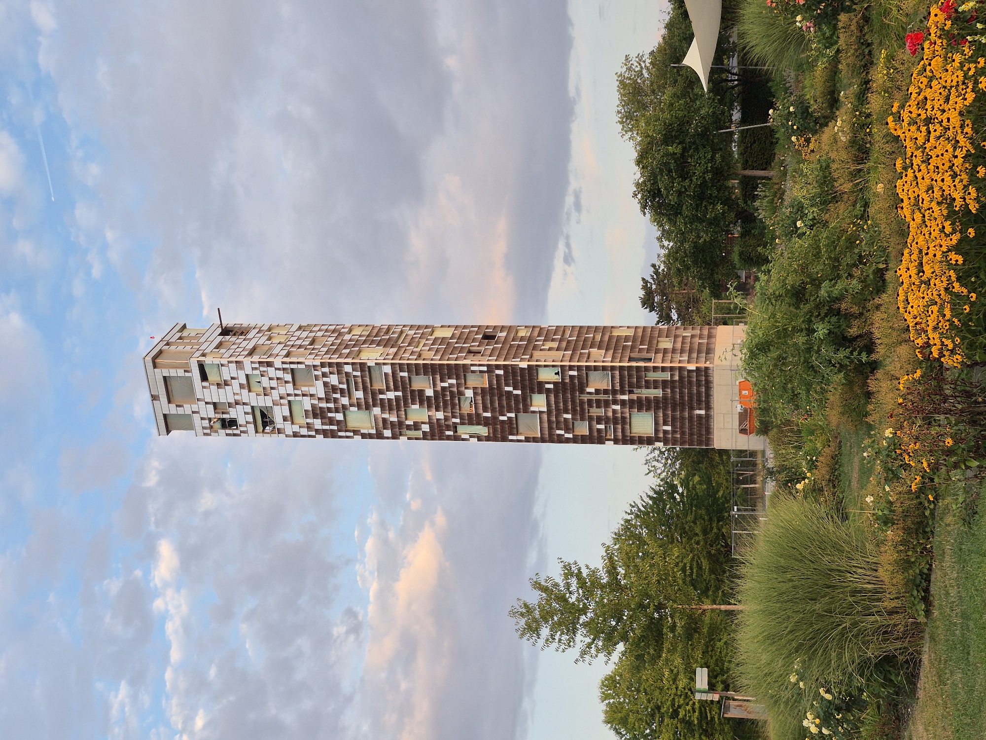 A tall, slender, contemporary tower stands prominently in the center of the frame, its facade a mosaic of brown and light-colored panels with numerous square and rectangular windows. The tower's upper section features a metallic finish. A person is visible in one of the windows near the top of the tower. The tower is situated in a verdant park-like setting. In the foreground, a lush garden is in bloom, with abundant yellow flowers, ornamental grasses, and other greenery. Behind the garden, trees line the middle ground, and a white, tent-like structure is partially visible on the right. The sky above is partly cloudy with hues of blue and soft pink, indicating either dawn or dusk. The scene is identified as the Remspark in Schwäbisch Gmünd, Germany.