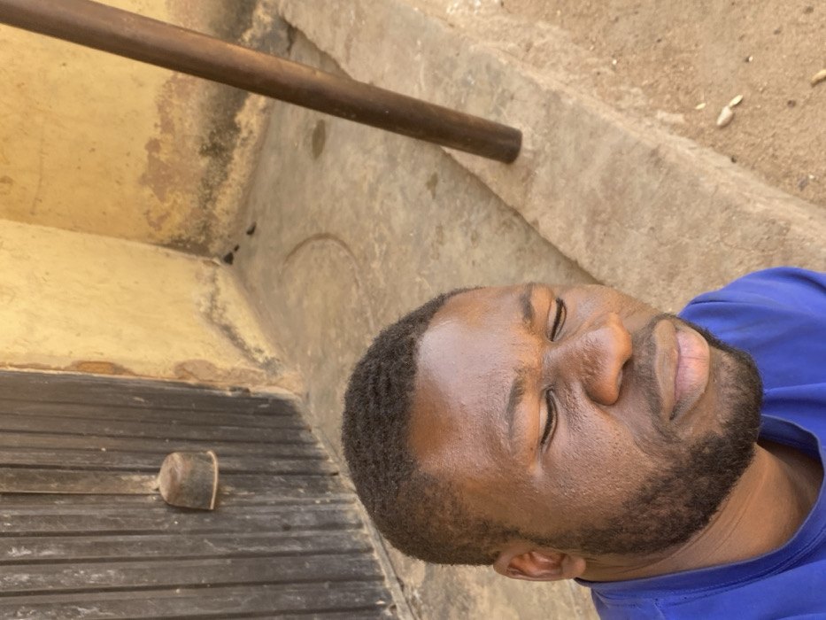 A dark-skinned male with a short beard and short, wavy dark hair, wearing a blue t-shirt, is depicted in a medium close-up, outdoors in Udu, Nigeria. His eyes are closed, and his face shows a slight grimace, suggesting discomfort or exposure to bright light. He is positioned in the lower-left foreground, looking downwards and to his right. The background features a weathered, light yellow-brown wall with dark stains and discoloration along its base. To the left, a dark, vertically paneled wooden door with a cylindrical, possibly metallic, fixture is visible. To the right, a dark brown vertical pole extends from the mid-ground upwards. The ground is a light-colored concrete or cement surface, showing a distinct semicircular groove and some small debris. The scene is illuminated by strong, direct sunlight.