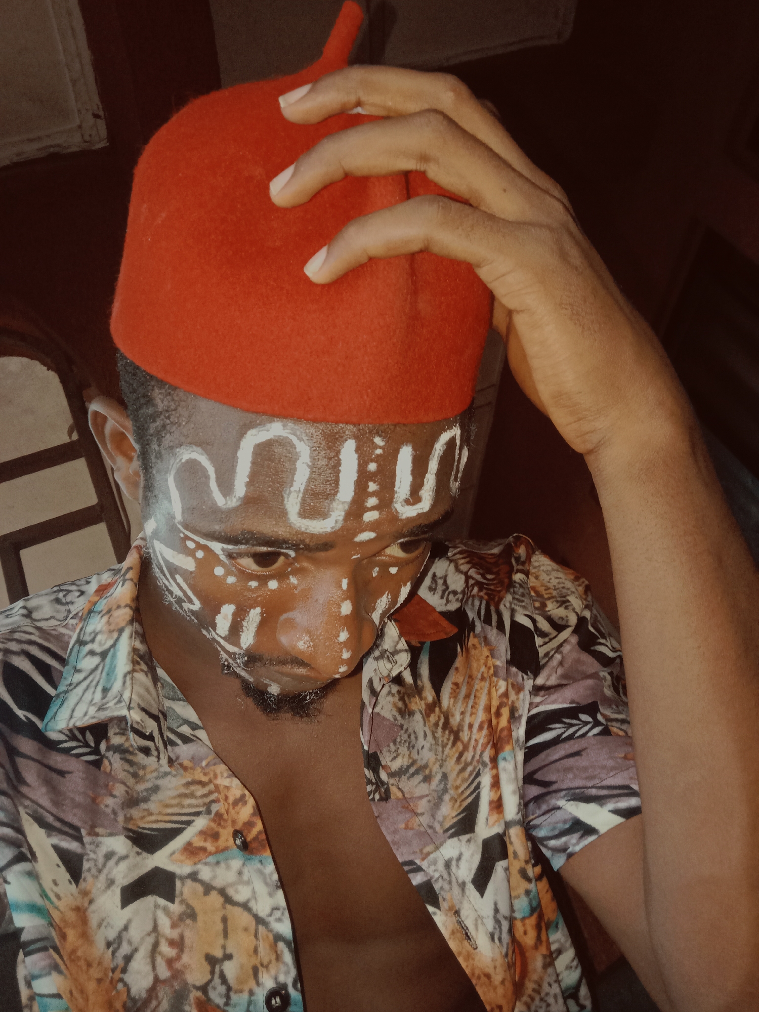 A close-up portrait features a person with dark skin, looking downwards with a pensive expression. They are wearing a vibrant red, traditional-style cap, which appears to be made of felt. One hand is gently placed on the red cap, with fingers visible. White traditional body paint or markings adorn the forehead in a distinct wavy pattern, and dots and lines are visible on the cheeks and around the chin, possibly for a cultural event or ritual. The person has short dark hair and a neatly trimmed beard. They are dressed in a patterned short-sleeved shirt with an intricate design featuring abstract and possibly natural motifs in shades of brown, beige, blue, and black. The shirt is unbuttoned at the collar, revealing a portion of the chest. The background is dimly lit and indistinct, suggesting an indoor setting or shaded area. No specific location cues or text are visible. The overall mood conveyed is serious or contemplative.