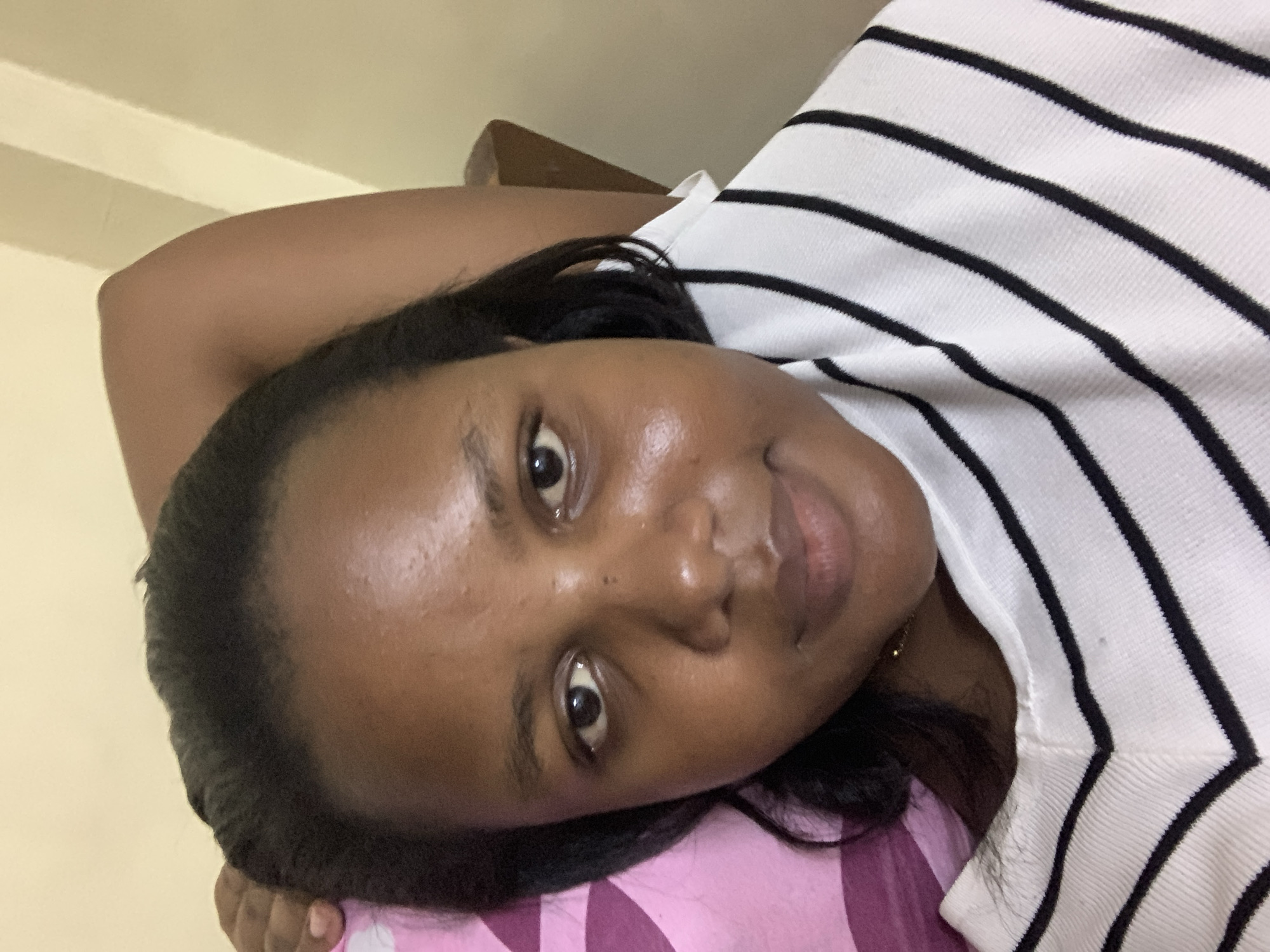 The image is a selfie of a dark-skinned woman with black hair, wearing a white and black striped shirt. She is lying down, with her head resting on a pink and purple patterned pillow or fabric. The background is a light yellow wall. There are no details indicating a location in Dar es-Salaam, Tanzania.