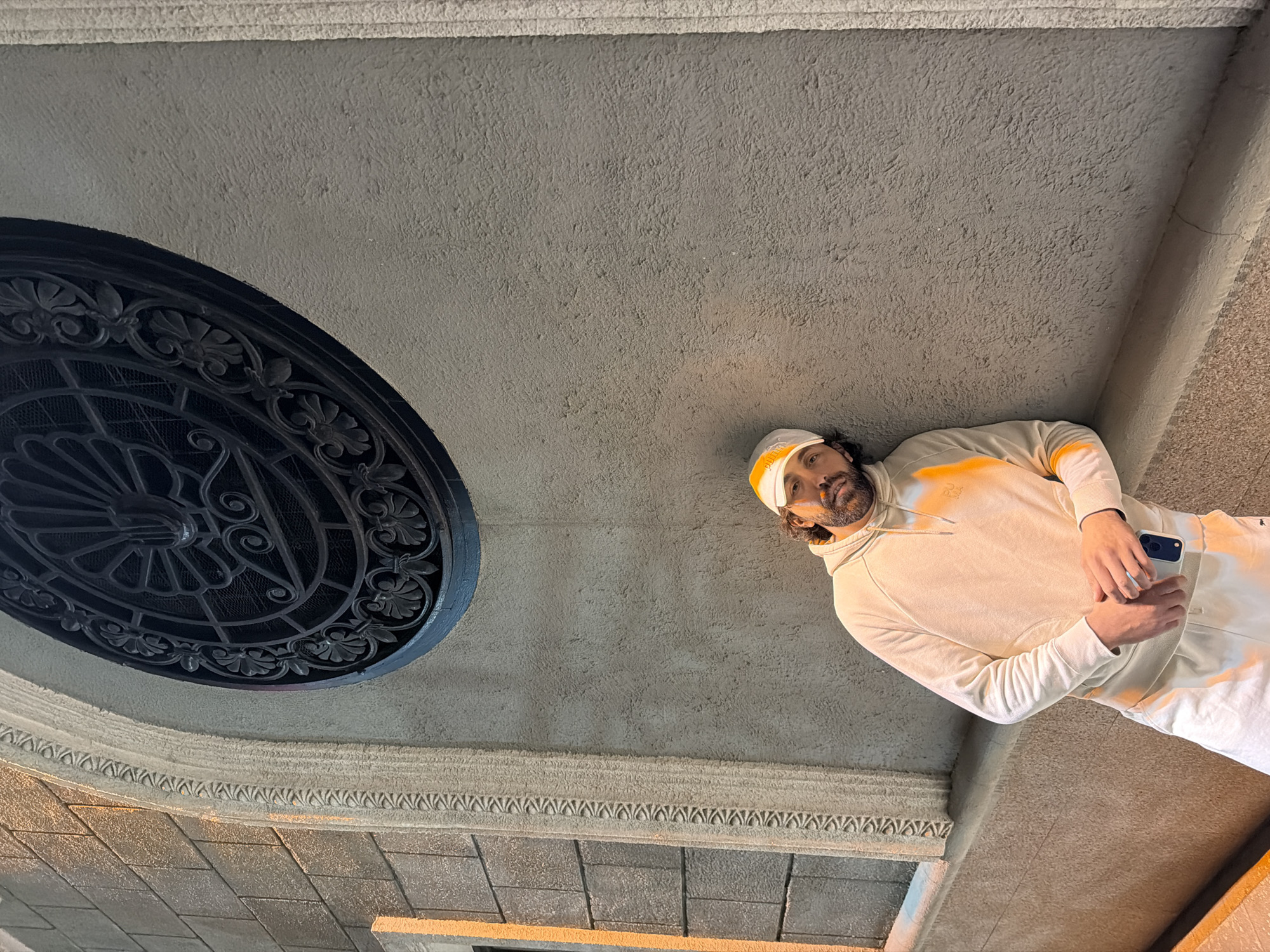 A man stands in the entry way of the Krasnoyarsk Regional Museum in Russia, beneath a decorative ceiling with an ornate, circular wrought iron design and patterned plasterwork. The man wears a white hoodie and matching sweatpants with orange details. He holds a smartphone in his hands.