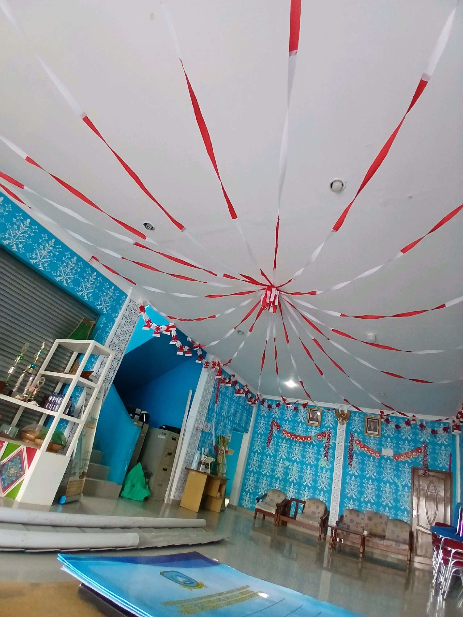 The image depicts an indoor space, possibly a community hall or school, decorated with red and white streamers hanging from the ceiling. The walls are adorned with blue wallpaper featuring a white snowflake pattern. In the foreground, a stack of blue folders or binders is visible on a table. To the left, a white shelf unit holds several trophies and some boxes. Further back, a staircase leads to a blue-painted upper level. In the main room, several pieces of furniture, including sofas and armchairs, are arranged. A wooden door is located on the right side of the room, with a stack of red chairs beside it. The overall impression is of a festive or celebratory atmosphere, possibly related to Indonesian Independence Day given the red and white decorations. The location is identified as Tarempa Timur, Indonesia. There are no people visible in the image.