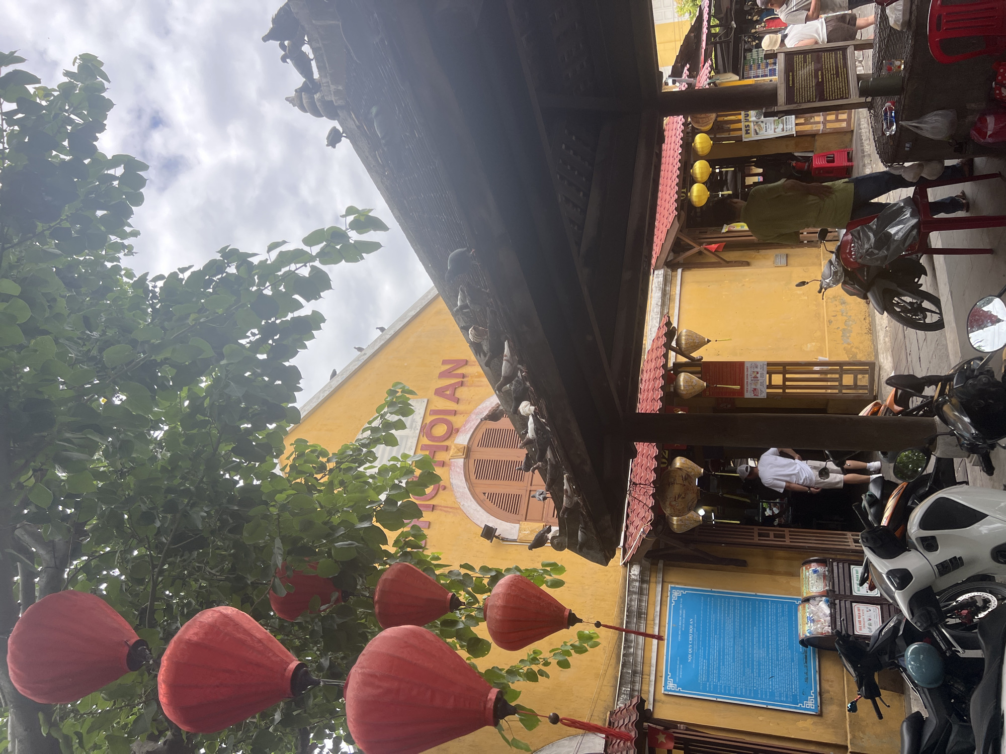 The image depicts a scene in Hoi An, Vietnam, likely within the historic Ancient Town, an area known for its vibrant yellow architecture, traditional lanterns, and bustling markets. Dominating the upper left of the frame are several large, red, paper lanterns hanging from the branches of a lush green tree. In the background, a bright yellow building with arched windows and the inscription "HOI AN" is visible.
Below the yellow building, a covered structure with a dark roof and red trim extends forward. This area appears to be an entrance to a market or shop. Several pigeons are perched on the roof of this structure, with more scattered around the roofline of the main building.
In the foreground, several motorcycles are parked. A man wearing a white t-shirt and beige shorts stands near the entrance of the covered structure, facing away from the camera with his hands on his hips. To the right, another man in a green and yellow striped shirt stands near a motorcycle, looking into the market. Various signs and posters are visible on the buildings, including a large blue sign with Vietnamese text to the left, likely containing market rules or information. Yellow paper lanterns are also hanging in the distance, adding to the festive atmosphere. The sky is overcast with scattered clouds.