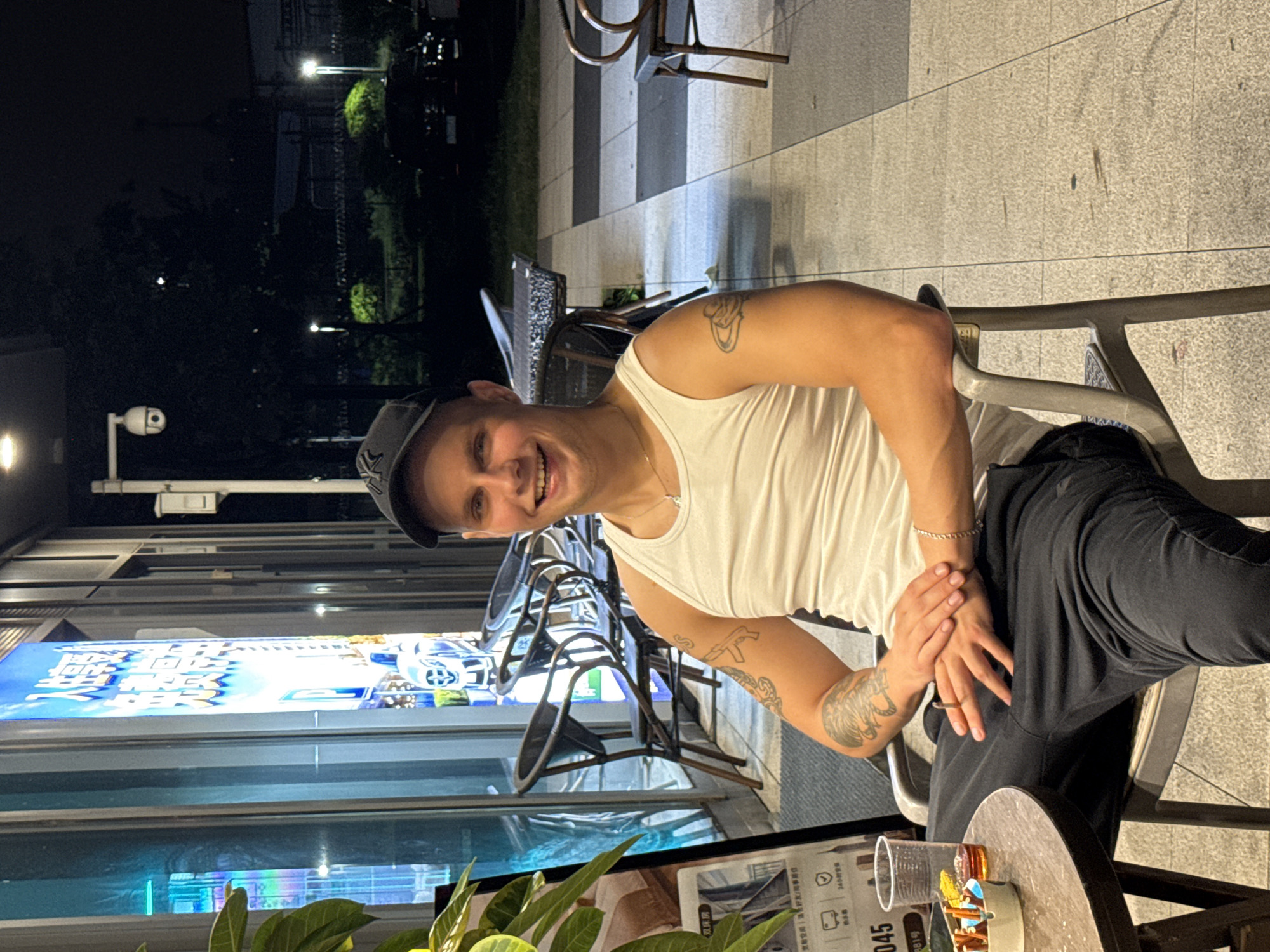 A man wearing a white tank top and a black baseball cap is sitting outdoors at night, with his legs crossed. He has tattoos on his arm and is smiling at the camera. A table with a drink and some colorful objects is in the foreground, and chairs are visible in the background. Behind the man, there is a large illuminated sign with what appears to be Chinese text and images of cars. The setting suggests an urban environment in Huangpu District, China, possibly a cafe or restaurant patio. The lighting indicates it is nighttime.