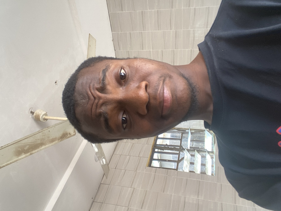 A close-up, head-and-shoulders indoor photograph depicts a dark-skinned male individual with short, dark hair, looking directly at the camera. His eyebrows are slightly furrowed, and his mouth is in a neutral or subtly smirking expression. He wears a black short-sleeved t-shirt. The background features a light-colored ceiling with a visible, discolored ceiling fan possessing at least two blades. The walls are covered with light-colored, horizontally arranged rectangular tiles. To the viewer's left, a window with multiple louvered glass panes and a metal frame offers a view of an indistinct exterior. This scene is situated within a room in Minna, Nigeria.