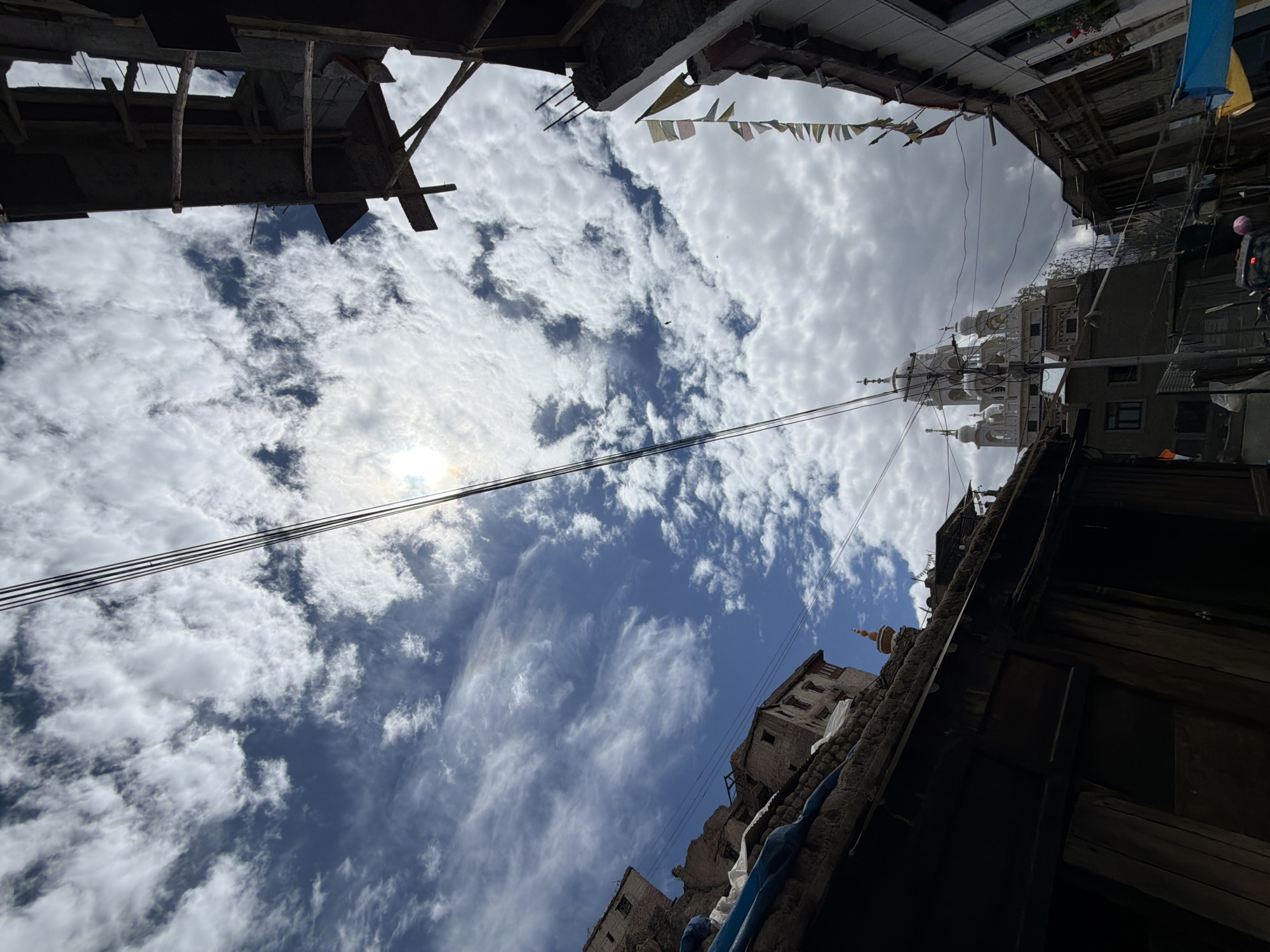 The image depicts a street scene in Leh, India, viewed from a low angle looking upwards. The sky is a prominent feature, filled with a mix of white, fluffy clouds and patches of blue. The sun is partially obscured by clouds, creating a bright, hazy effect. Power lines run diagonally across the frame, adding a sense of urban infrastructure.
To the left, traditional stone buildings with wooden elements are visible, some appearing aged and weathered. A blue tarp is draped over a section of one of these buildings. In the distance, a large, ornate white structure with multiple domes and spires stands out against the sky, suggesting it might be a monastery or a temple. Prayer flags, colorful and rectangular, are strung along the right side of the frame, attached to buildings that appear to be under construction or in a state of disrepair, with visible wooden beams and concrete.
The foreground is dominated by the dark interior of a building or overhang, with rough-hewn wooden planks and beams. A small, pointed spire of a building is visible between the foreground structure and the white domed building. The overall impression is of a busy, perhaps somewhat rustic, urban environment with a blend of traditional architecture and functional infrastructure, set against a dramatic sky.