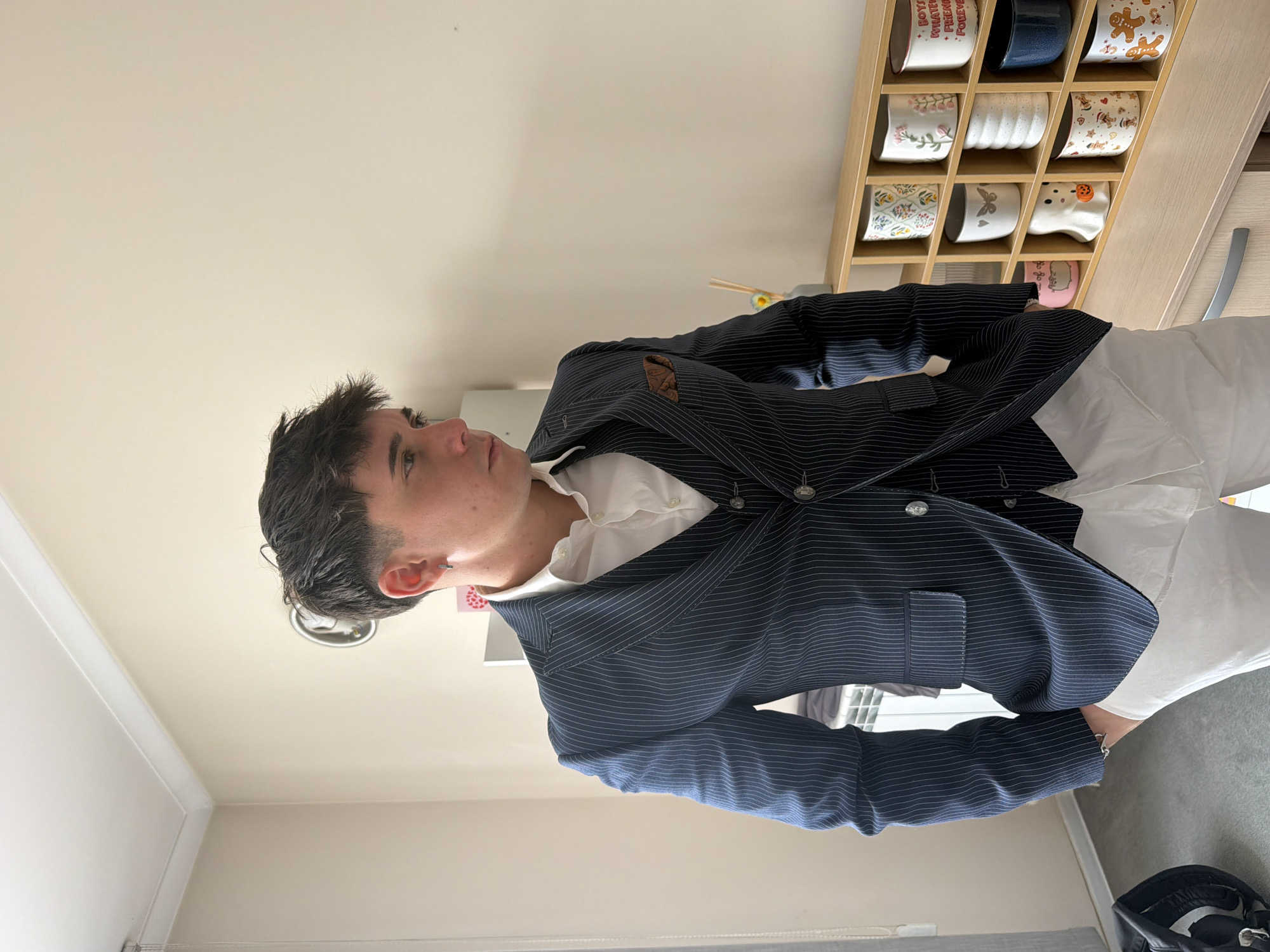 A young man with dark hair is wearing a navy pinstripe suit with a white shirt and white pants. The suit jacket is buttoned, and he has a pocket square. He is standing indoors, with a shelf of mugs visible in the background. A ceiling light and a small portion of a window are also visible. The exact location within Sofia, Bulgaria, cannot be determined from the provided image.