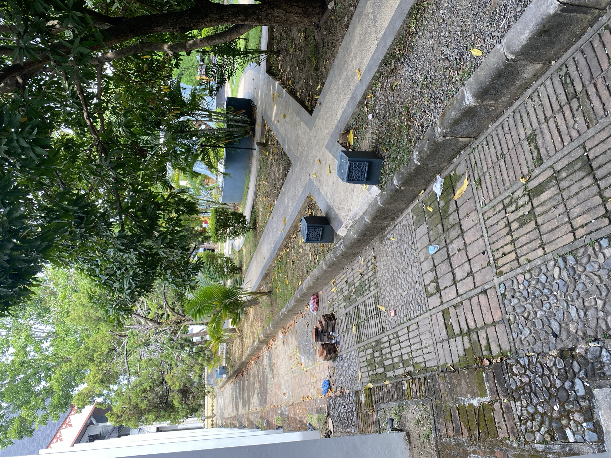 The image depicts an outdoor pathway in a green space, possibly a park or campus, in Banda Aceh, Indonesia. A white building with decorative trim is visible on the left. The pathway is constructed with a mix of red bricks and grey stones, with some areas showing moss and wear. Along the edge of the pathway, there is a concrete curb bordering a gravelly area with scattered leaves and some vegetation.
Further along the pathway, there is a section with a concrete walkway that diverges into multiple paths, forming a cross shape. Two dark grey, square planters with intricate cutouts are placed near the junction of these concrete paths. Surrounding the concrete walkways are areas of grass and soil, with several palm trees and other lush green foliage present. In the background, a light blue curved structure and a distant orange vehicle or piece of equipment can be observed.
On the brick and stone pathway in the foreground, there are several objects: a small stack of red bricks, a clear plastic bag with red contents, and two small blue plastic bags. Scattered leaves are present on the pathways. The environment appears to be a well-maintained outdoor area, albeit with some signs of weathering and litter.